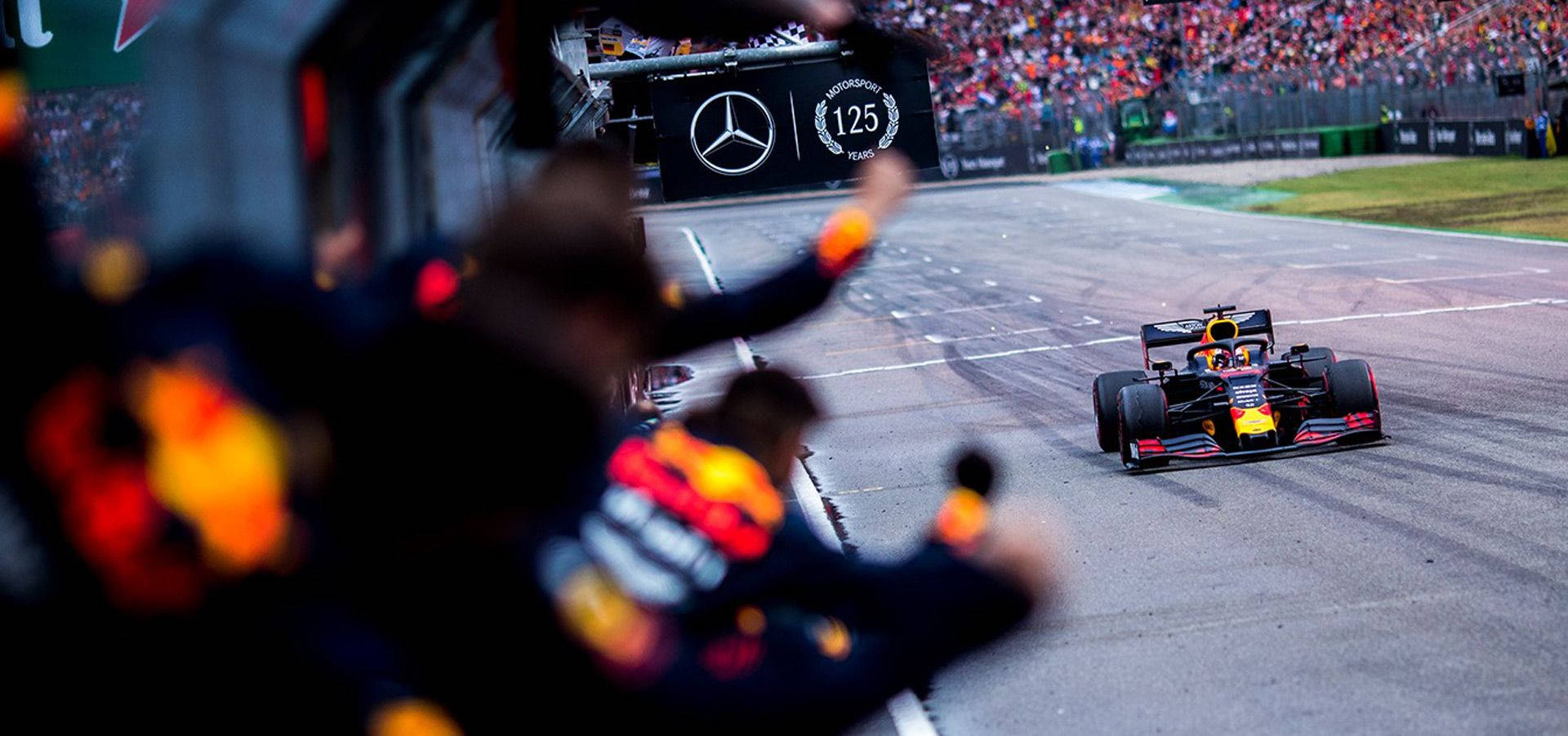 Verstappen 桌布 壁紙 — 一輛紅牛一級方程式賽車高速駛過在賓士125周年紀念橫幅前揮手的維修站人員。
