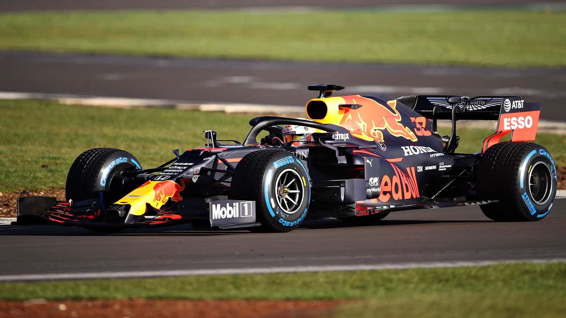 Max Verstappen 桌布 壁紙 — 一輛紅牛車隊一級方程式賽車在賽道上疾馳，車身採用鮮豔的紅、黃、黑色塗裝，背景是綠色草地和瀝青路面。