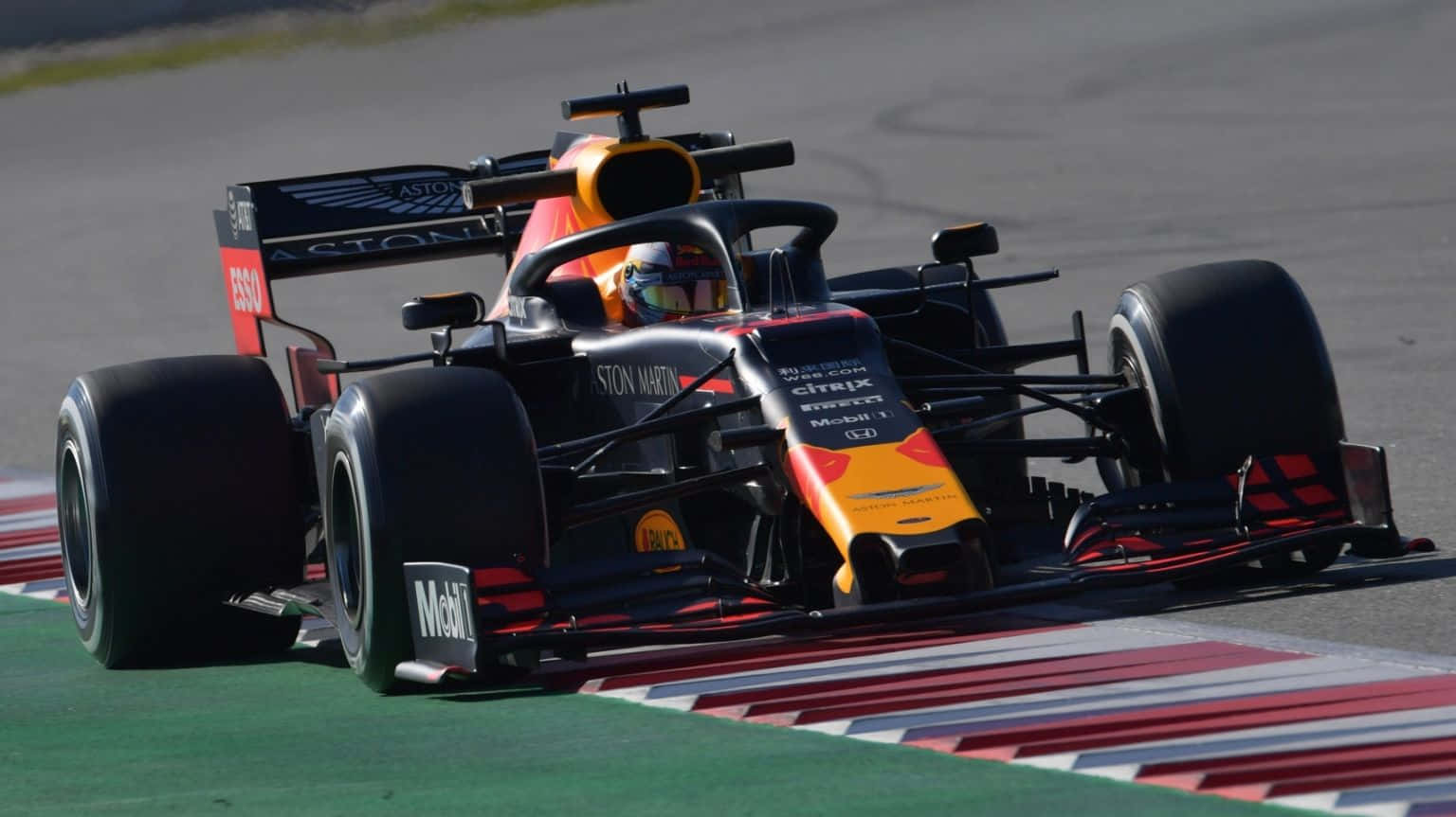 Max Verstappen 桌布 壁紙 — 一輛紅牛車隊一級方程式賽車在陽光普照下，沿著設有紅、白、綠色路緣石的賽道奔馳。