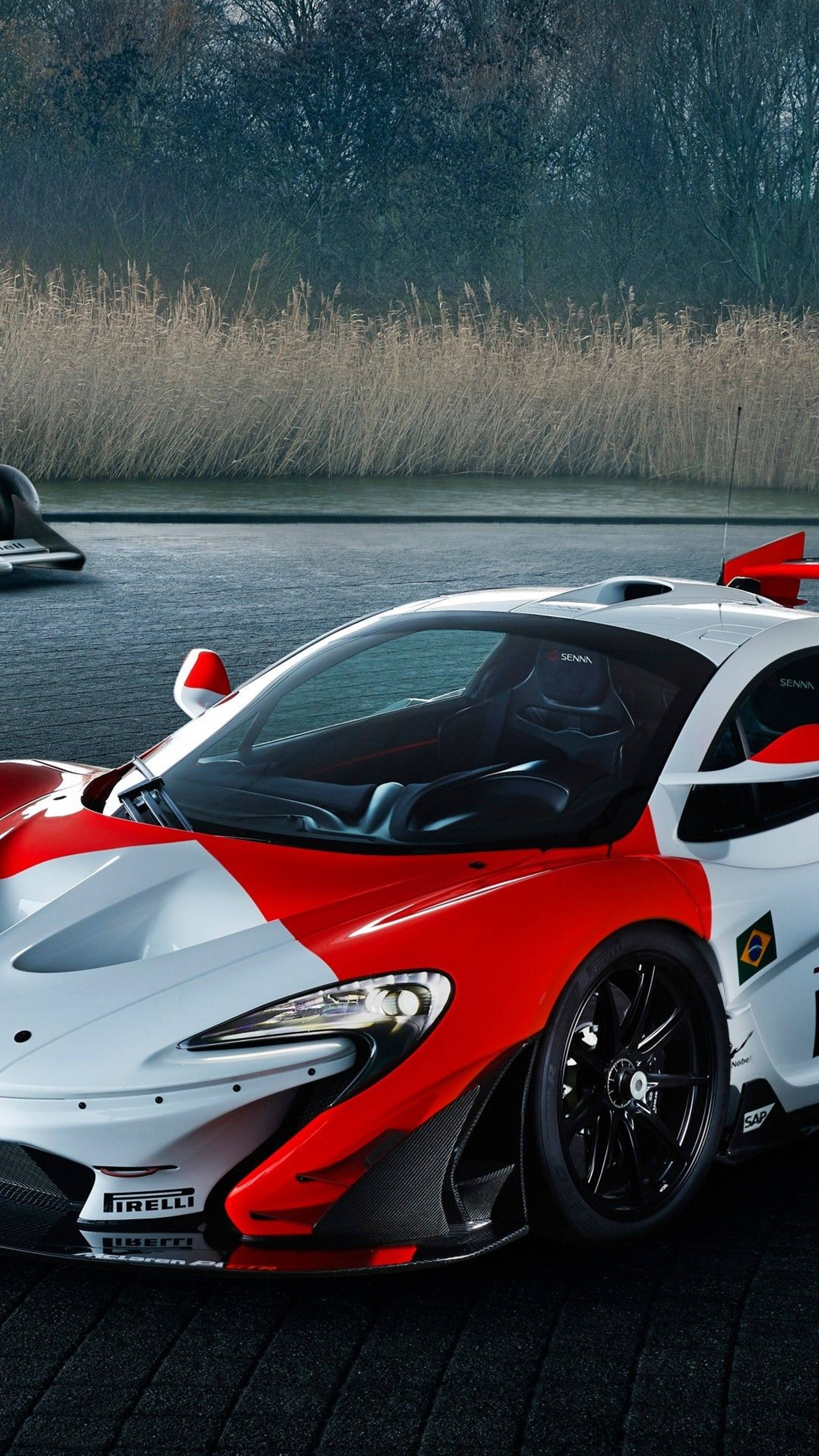 Luxury 桌布 壁紙 — 一輛紅白相間的McLaren Senna賽車停放在深色鵝卵石路面上，旁邊是一條平靜的水道，背景有蘆葦和樹木。