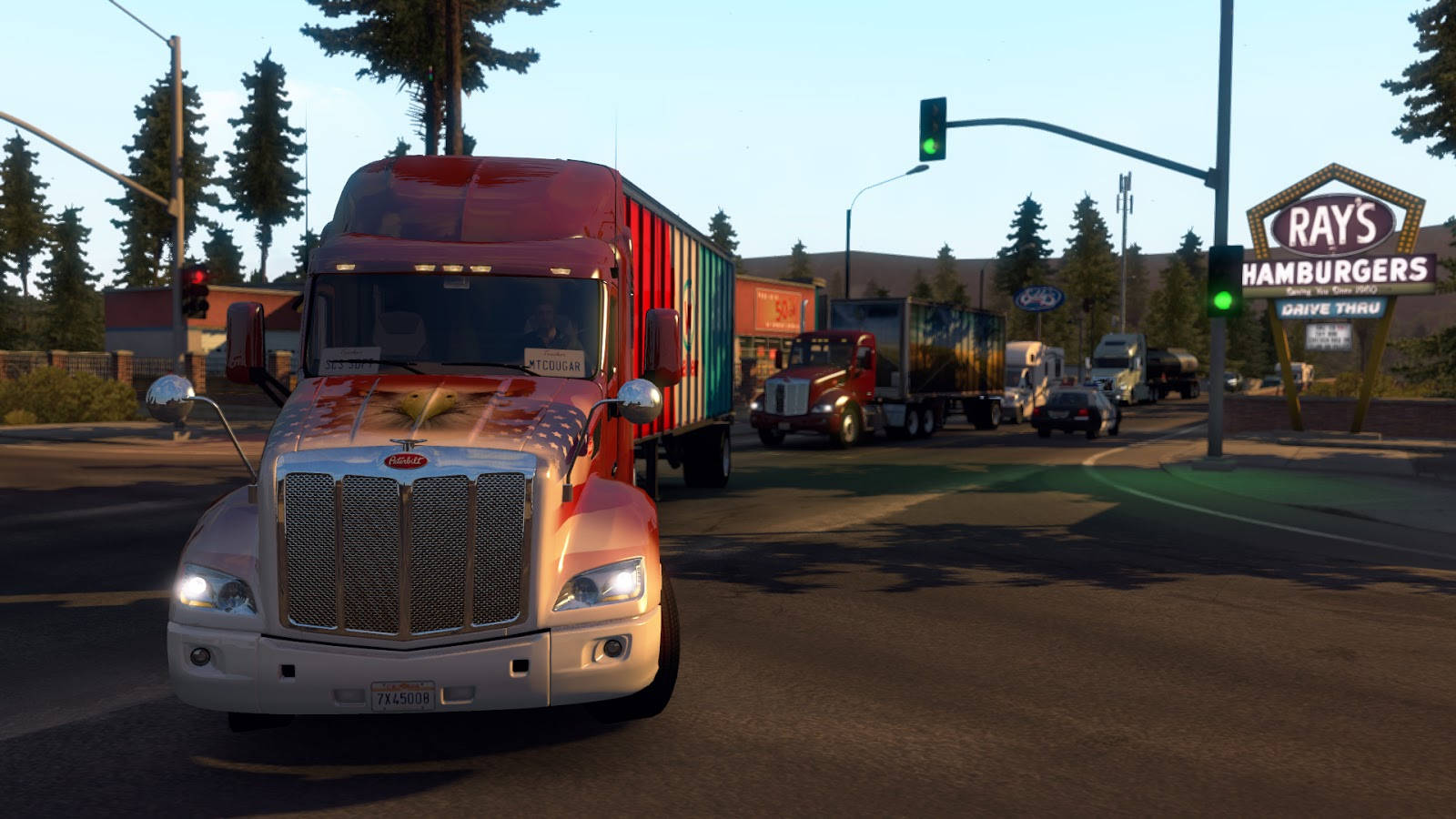 American Truck Simulator 桌布 壁紙 — 一輛紅白相間的半掛卡車拖著色彩繽紛的拖車，在晴朗的天空下於Ray's Hamburgers附近的交通燈前等待。