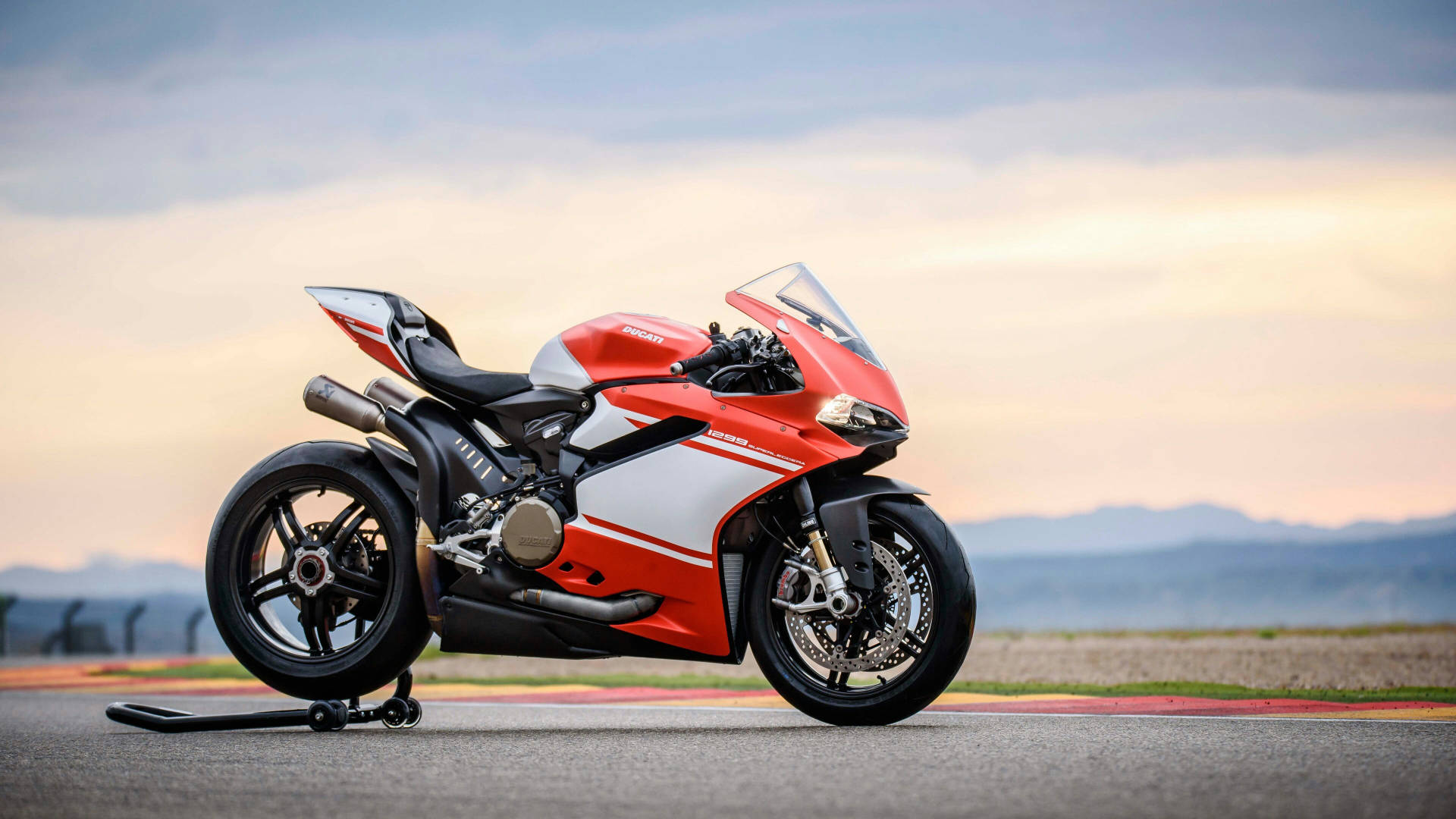 杜卡迪 桌布 壁紙 — 一輛紅白色 Ducati 摩托車在日落時分的賽道上蓄勢待發，遠山在柔和天空下。