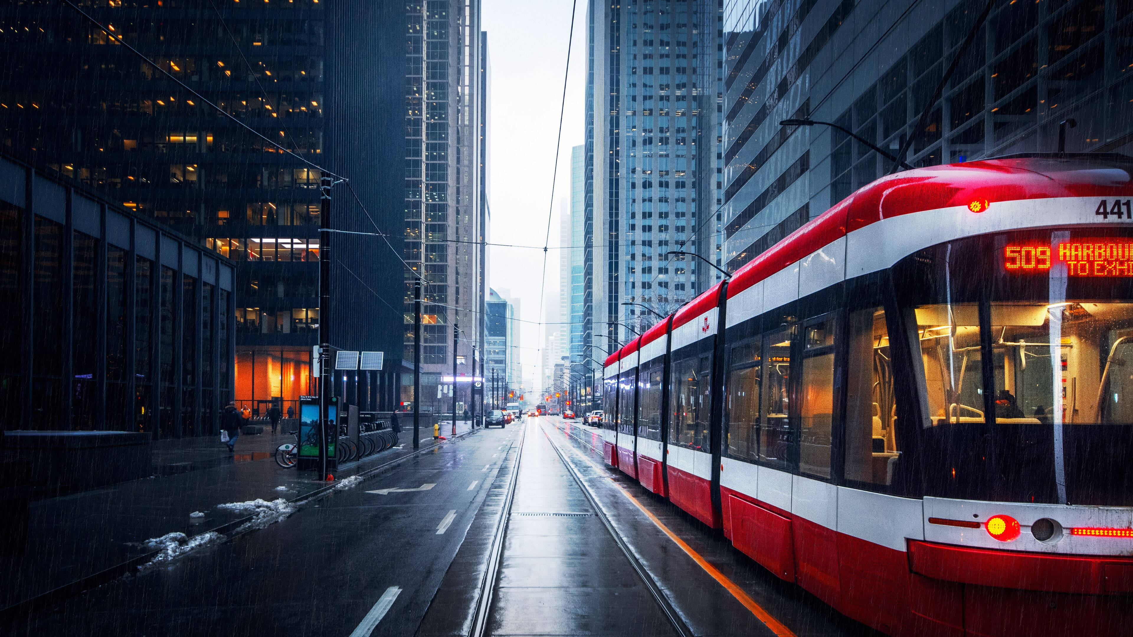 Toronto 桌布 壁紙 — 一輛紅白色的電車在灰濛濛的天空下，沿著被雨水浸濕的都市峽谷滑行，兩旁是高聳的玻璃摩天大樓。