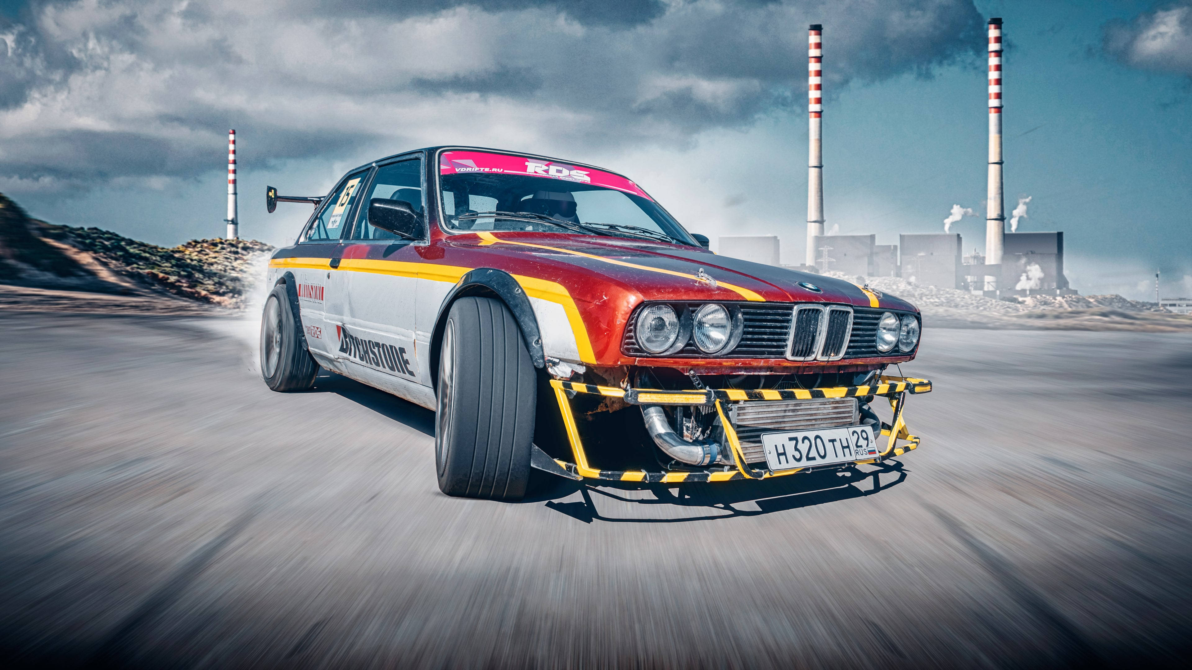 Clouds Aesthetics 桌布 壁紙 — 一輛紅白黃色的BMW賽車在多雲的天空下，以工業煙囪為背景，在塵土飛揚的賽道上飛馳。