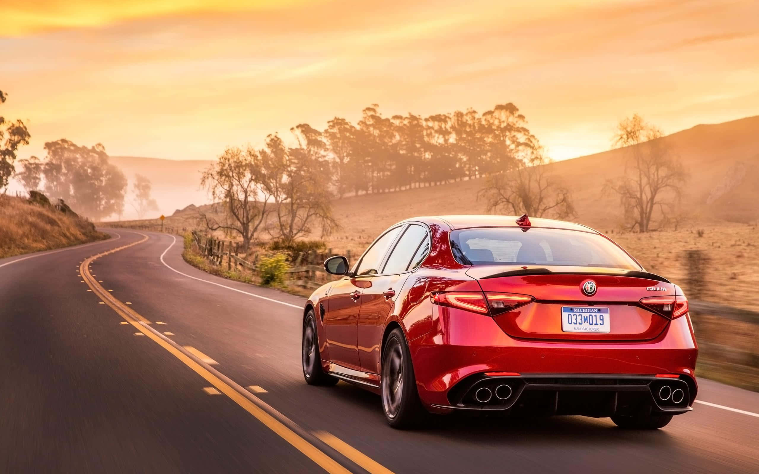 Alfa Romeo Giulia 桌布 壁紙 — 一輛紅色 Alfa Romeo 房車在金色夕陽下，沿著蜿蜒的鄉村道路飛馳，背景是山丘和樹木。
