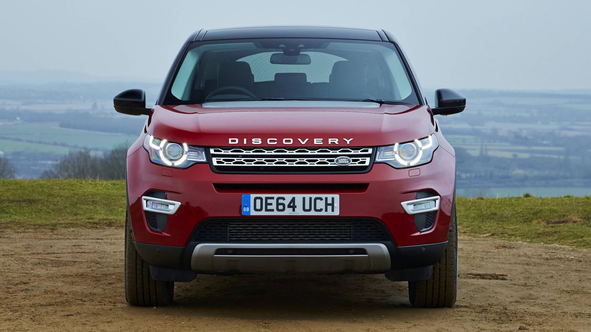 land rover iPhone 桌布 壁紙 — 一輛紅色 Land Rover Discovery 正面朝前停在泥路上，俯瞰著陰天籠罩下霧氣瀰漫的鄉村山谷。