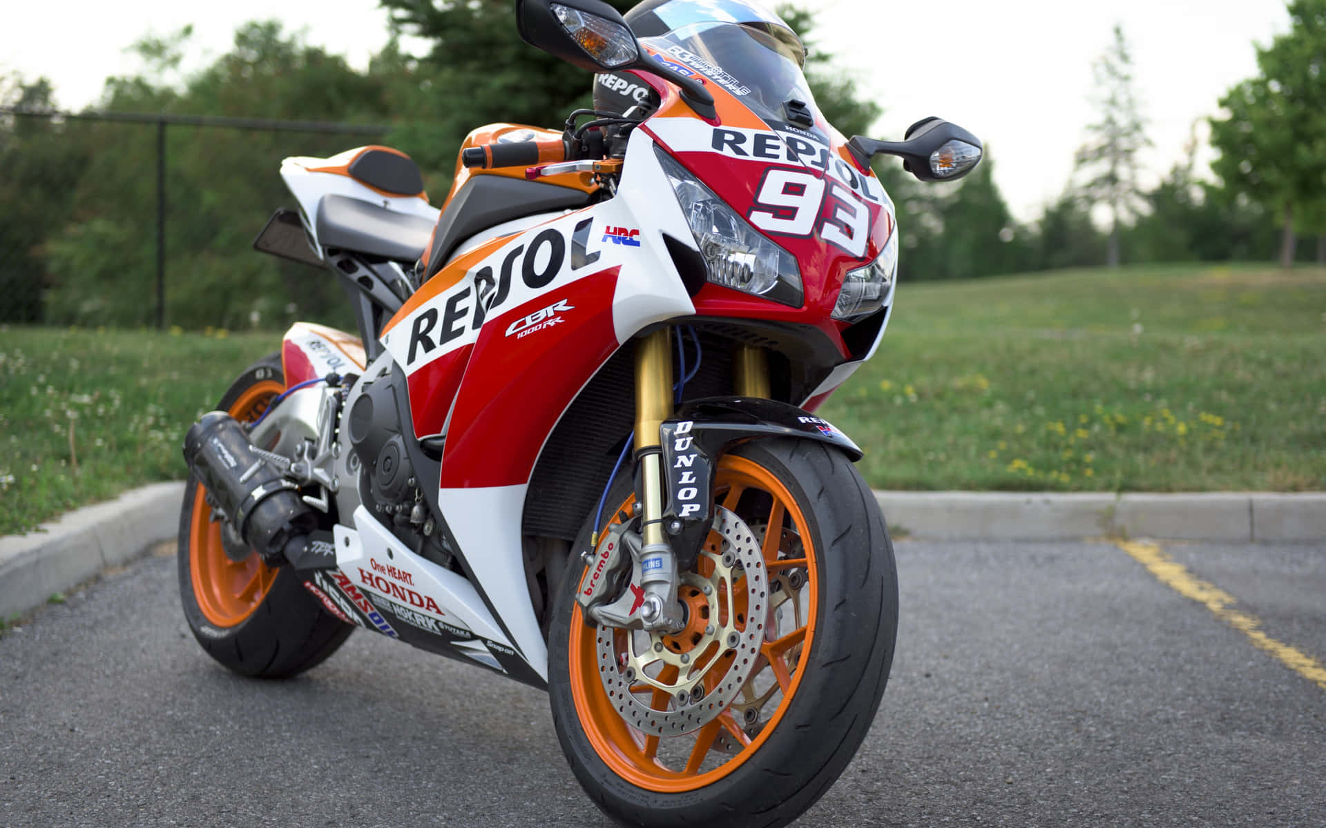  壁紙 — 一輛紅色、白色和橙色的Repsol Honda CBR賽車機車停在柏油路上，背景有綠草和樹木。