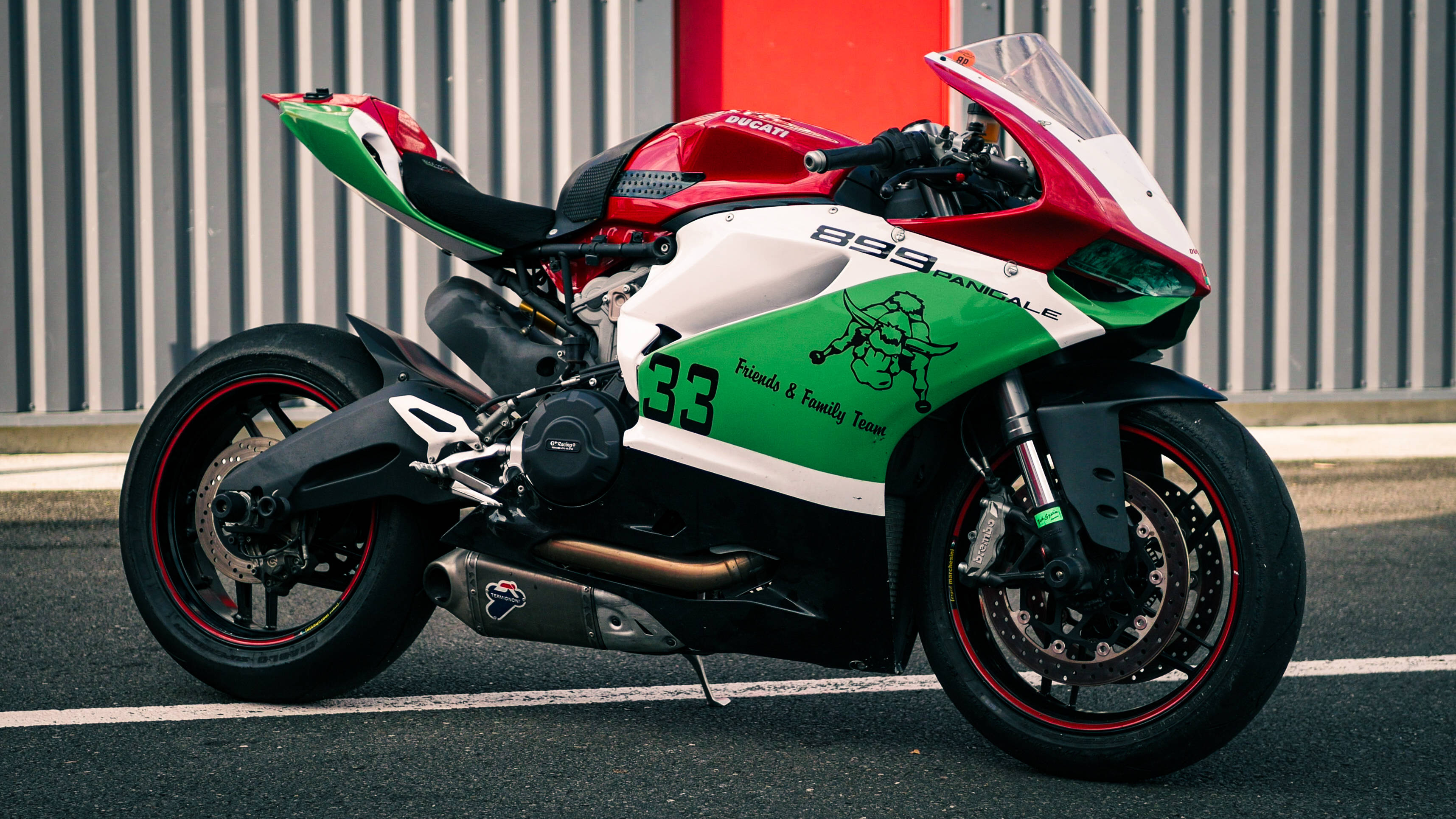 杜卡迪 桌布 壁紙 — 一輛紅色、白色和綠色的Ducati Panigale摩托車，編號33，停在瀝青路上，靠著波紋金屬牆。