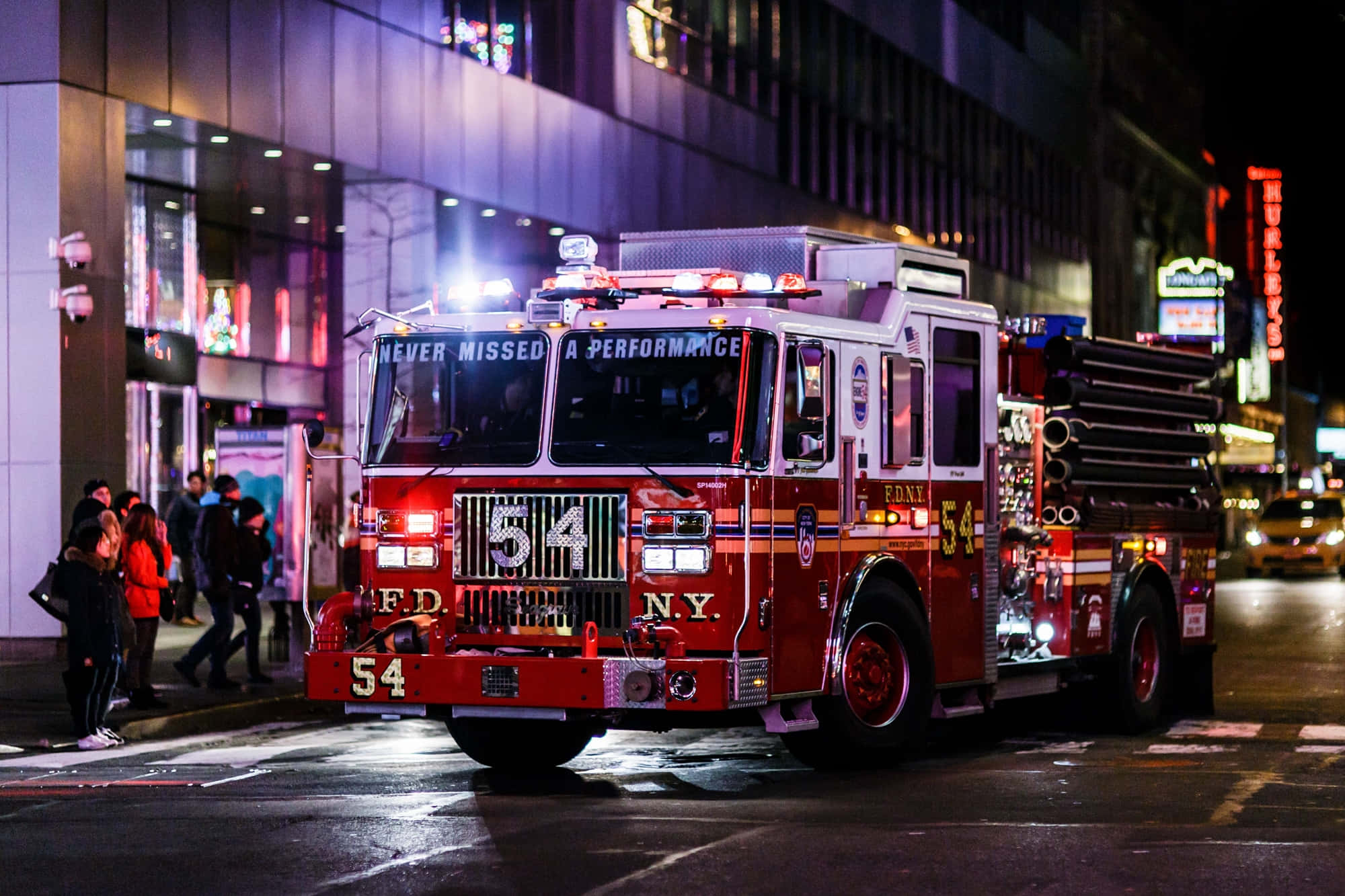 Fire Truck 桌布 壁紙 — 一輛紅色FDNY消防車閃著燈停在夜晚濕漉漉的城市街道上，周圍有行人和發光的霓虹燈招牌。