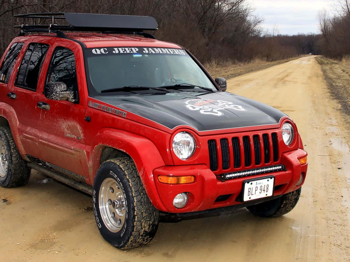 Jeep Liberty 桌布 壁紙 — 一輛紅色Jeep Liberty，黑色引擎蓋和車頂行李架，停放在泥濘的土路上，周圍是光禿禿的樹木。