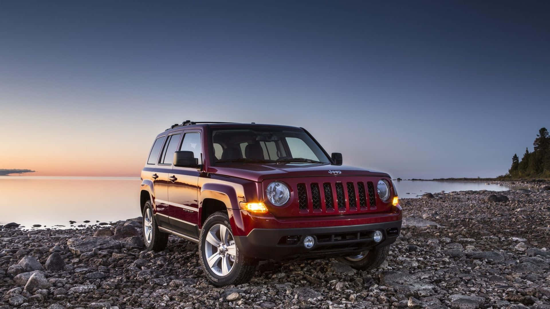 Jeep Patriot 桌布 壁紙 — 一輛紅色Jeep Patriot在日落時停泊在岩石湖岸，平靜的水面和遠處的樹木。
