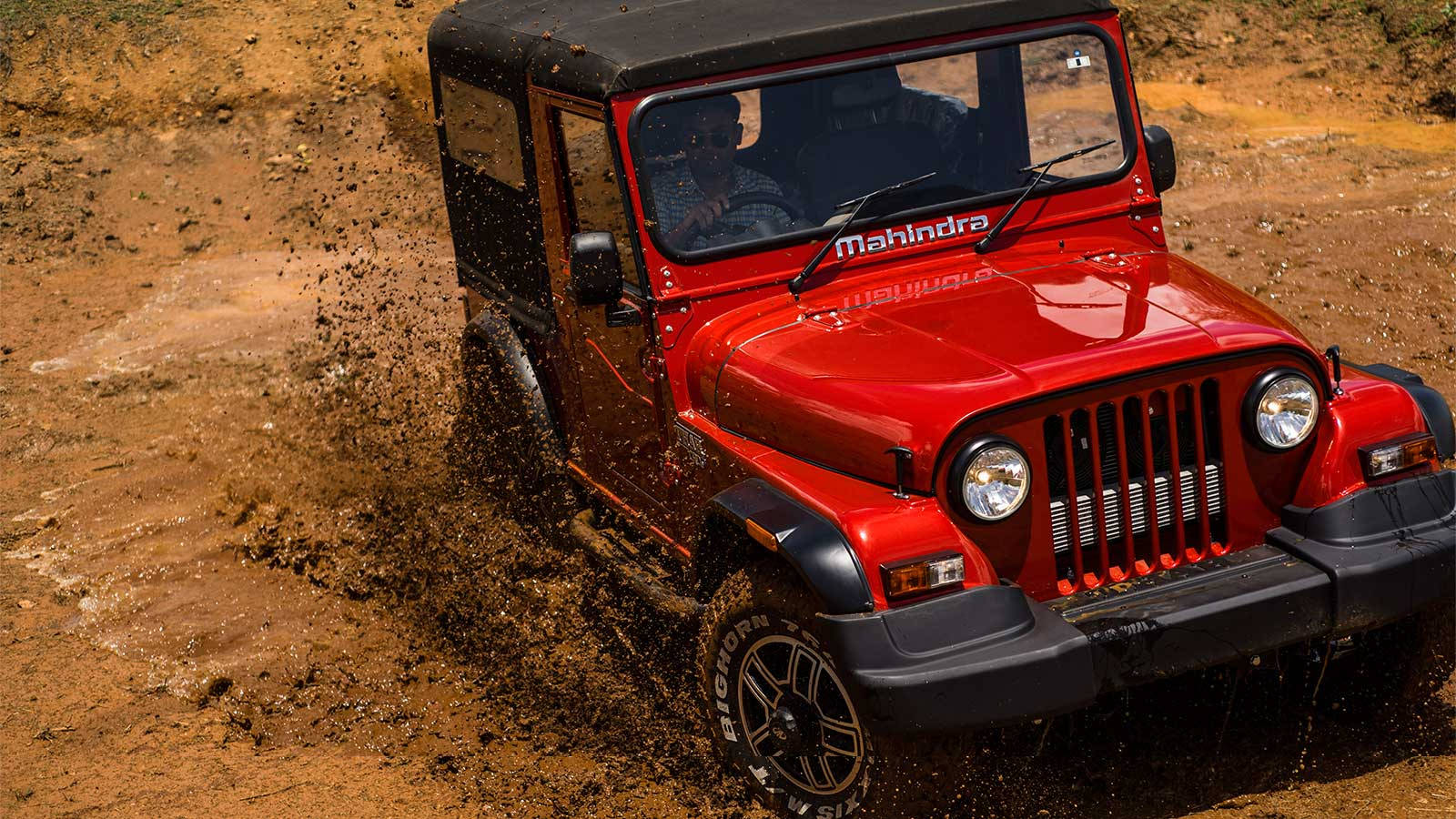 高清 桌布 壁紙 — 一輛紅色Mahindra Thar吉普車在泥濘地形中強勁行駛，在崎嶇的戶外環境中揚起泥土。