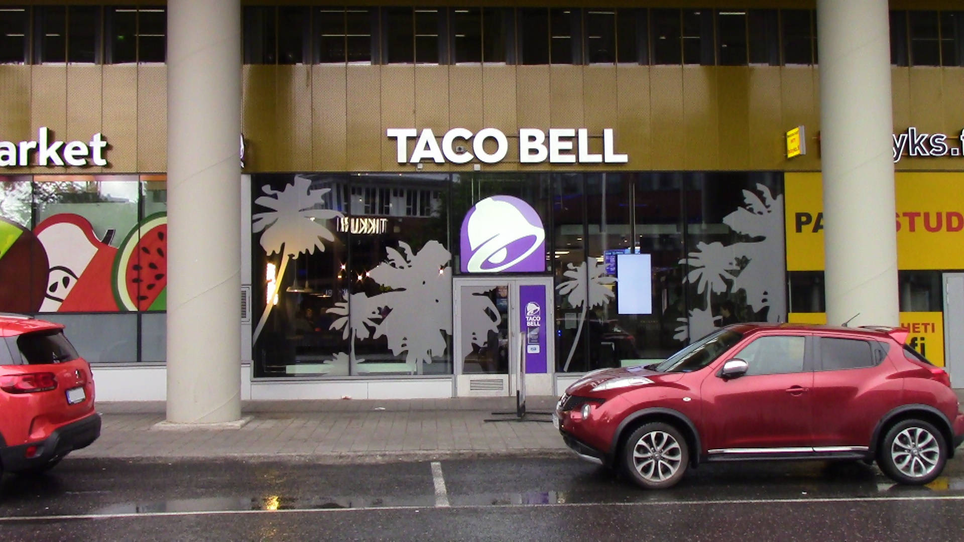塔可鐘 桌布 壁紙 — 一輛紅色SUV停在Taco Bell餐廳前，玻璃窗上有棕櫚樹貼紙，濕漉漉的人行道反射著城市燈光。