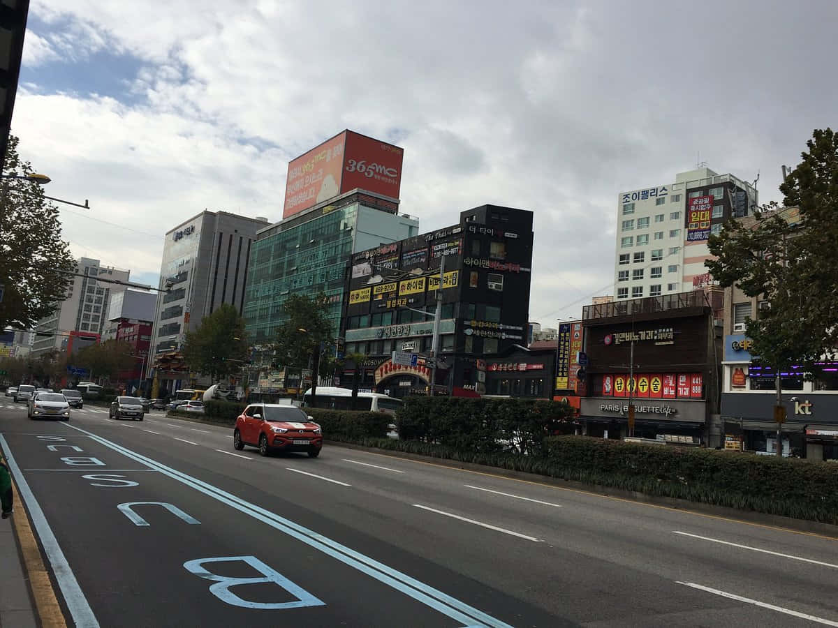 Street 桌布 壁紙 — 一輛紅色SUV在陰天下行駛在一條繁忙的城市街道上，街道兩旁是現代建築和色彩繽紛的韓文招牌。