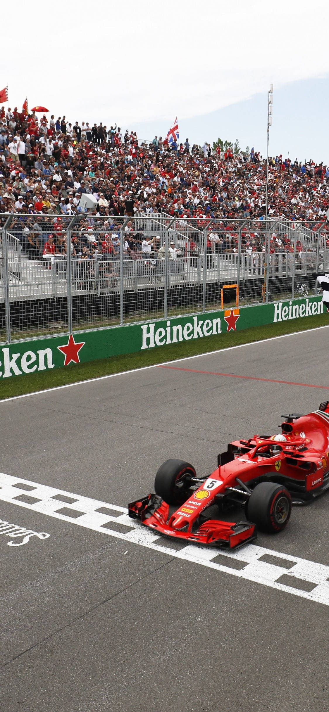 F1 iPhone 壁紙 桌布 壁紙 — 一輛紅色一級方程式賽車越過方格旗終點線，安全屏障後的看台上擠滿了歡呼的車迷。