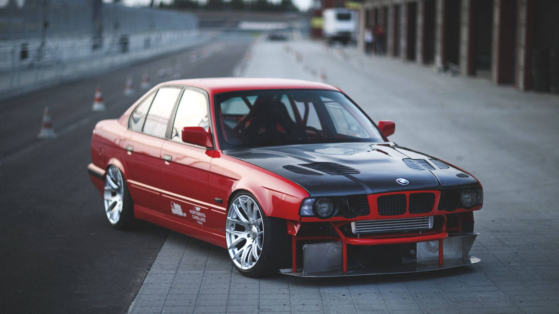 Road 桌布 壁紙 — 一輛紅色和黑色改裝的BMW E36轎車，配備自訂寬體套件和賽車輪，停放在鋪砌的賽道旁。