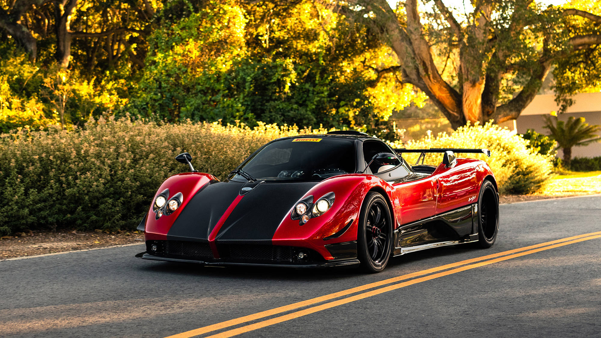 cars 桌布 壁紙 — 一輛紅色和黑色的Pagani Zonda跑車沿著陽光明媚的道路行駛，兩旁是金色的樹木和綠色的籬笆。