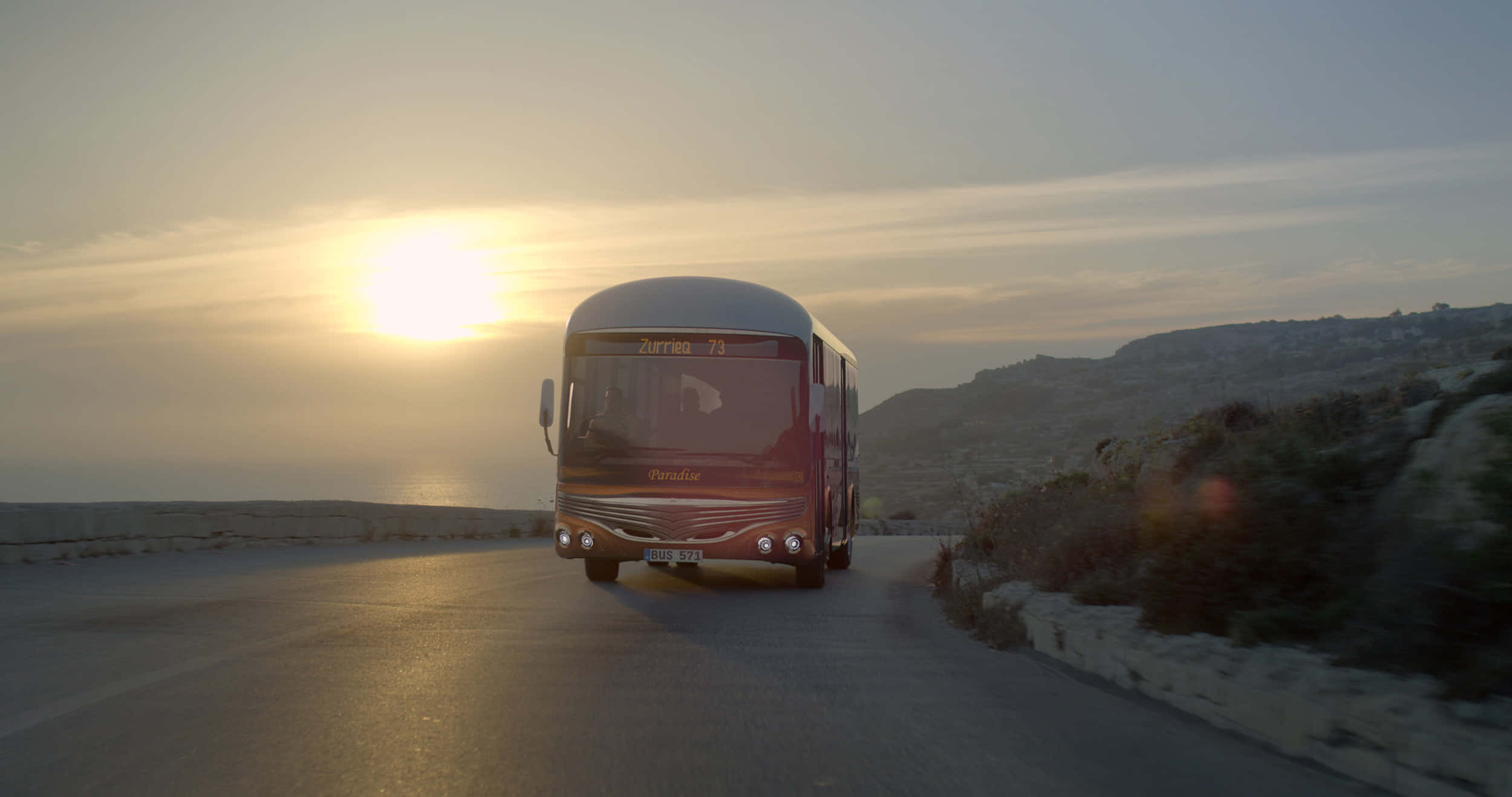 Bus 桌布 壁紙 — 一輛紅色巴士在海濱公路上駛向鏡頭，夕陽在海面上灑下金色光芒。