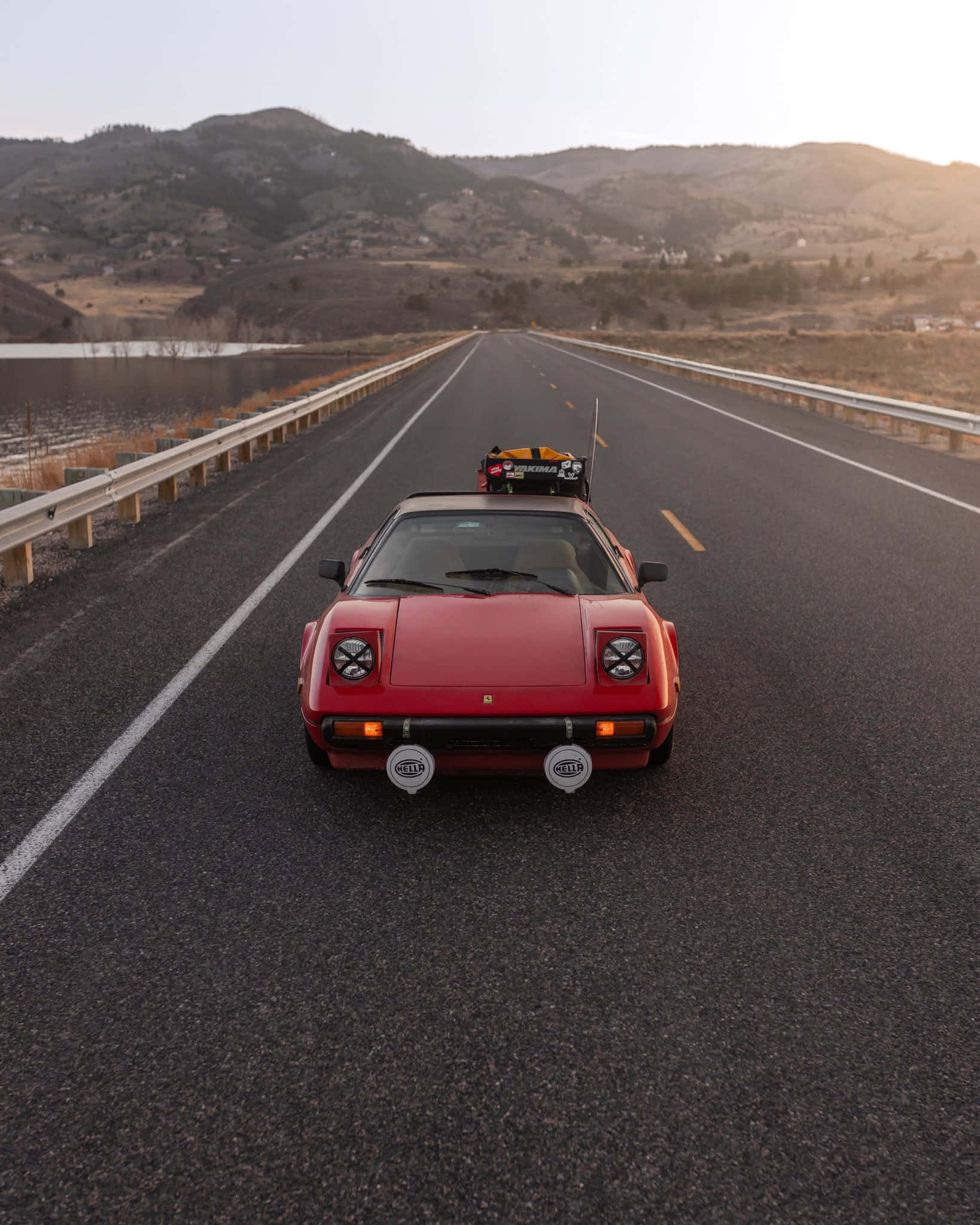Vintage Ferrari 桌布 壁紙 — 一輛紅色復古跑車，車頂放著賽車頭盔，在黃金時段沿著一條兩旁有山丘的長直路行駛。