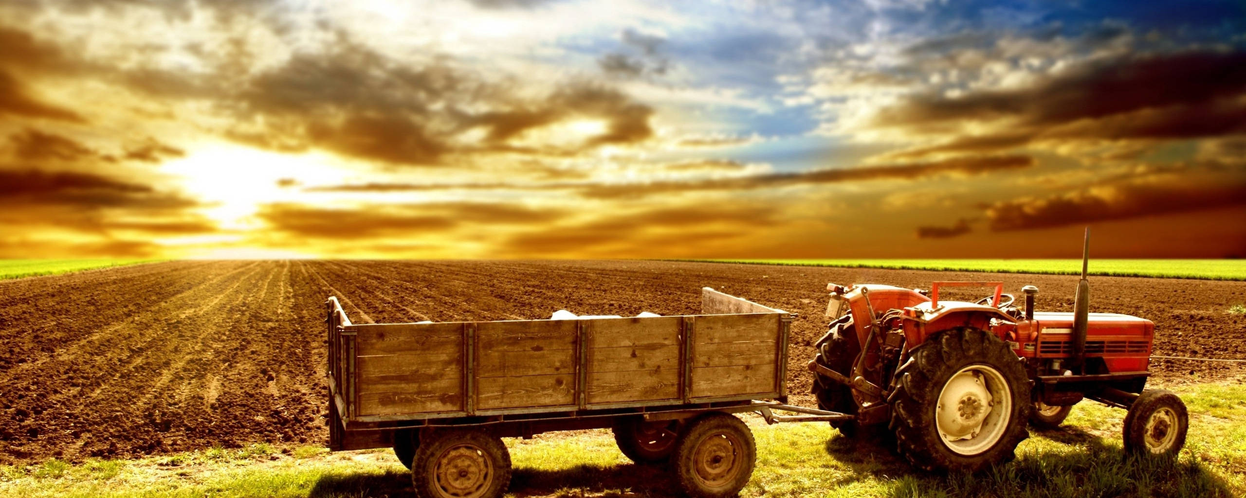 Agriculture 桌布 壁紙 — 一輛紅色拖拉機拖著木製馬車，在戲劇性的金色日落天空下穿過剛犁過的田地。