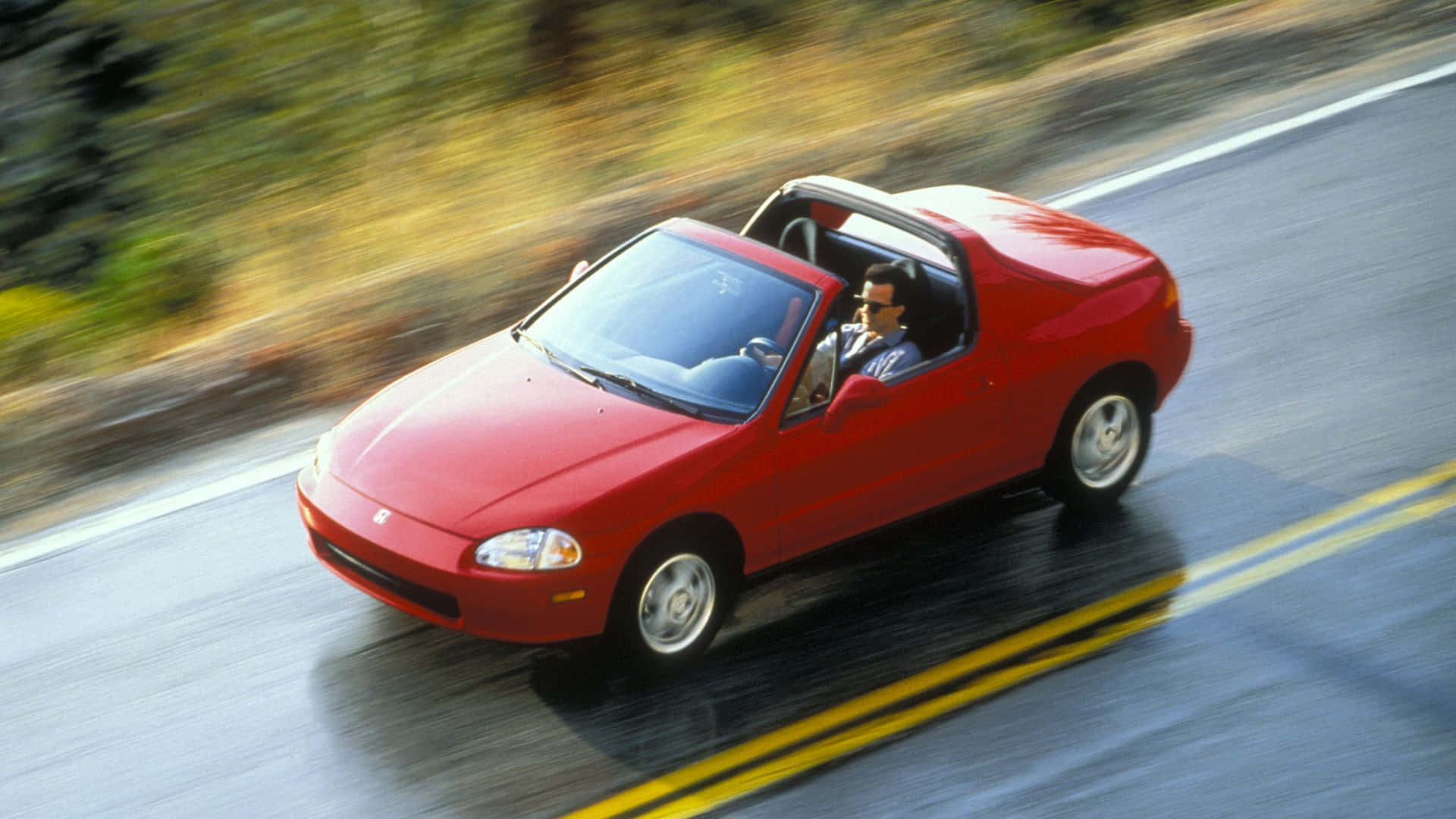 Honda Civic Del Sol 桌布 壁紙 — 一輛紅色本田Miata敞篷跑車在蜿蜒的道路上疾馳，周圍是模糊的綠化和黃線。