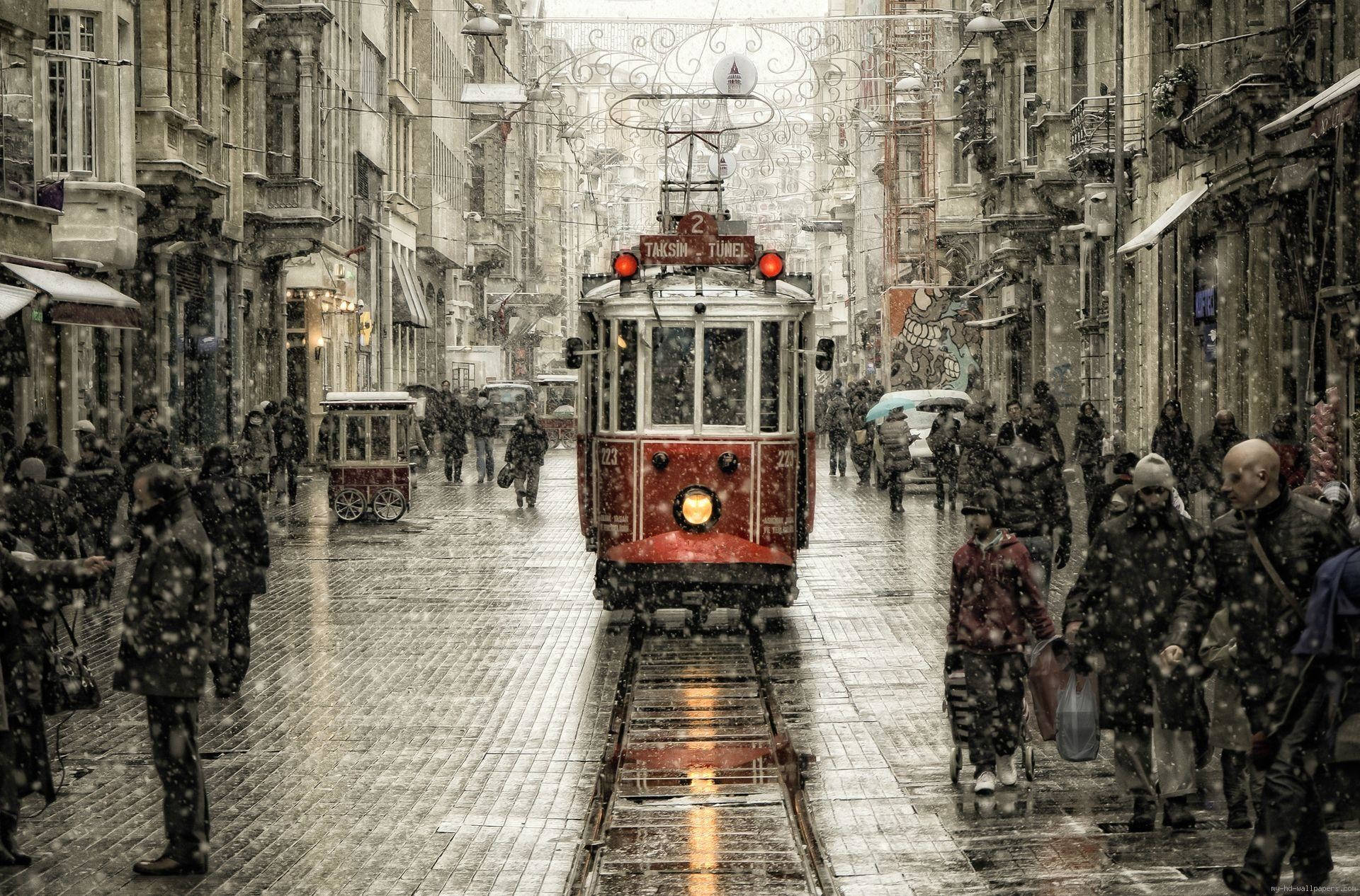 伊斯坦堡 桌布 壁紙 — 一輛紅色歷史電車在積雪的歐洲街道上滑行，行人則在飄雪中行走在濕滑的鵝卵石路上。