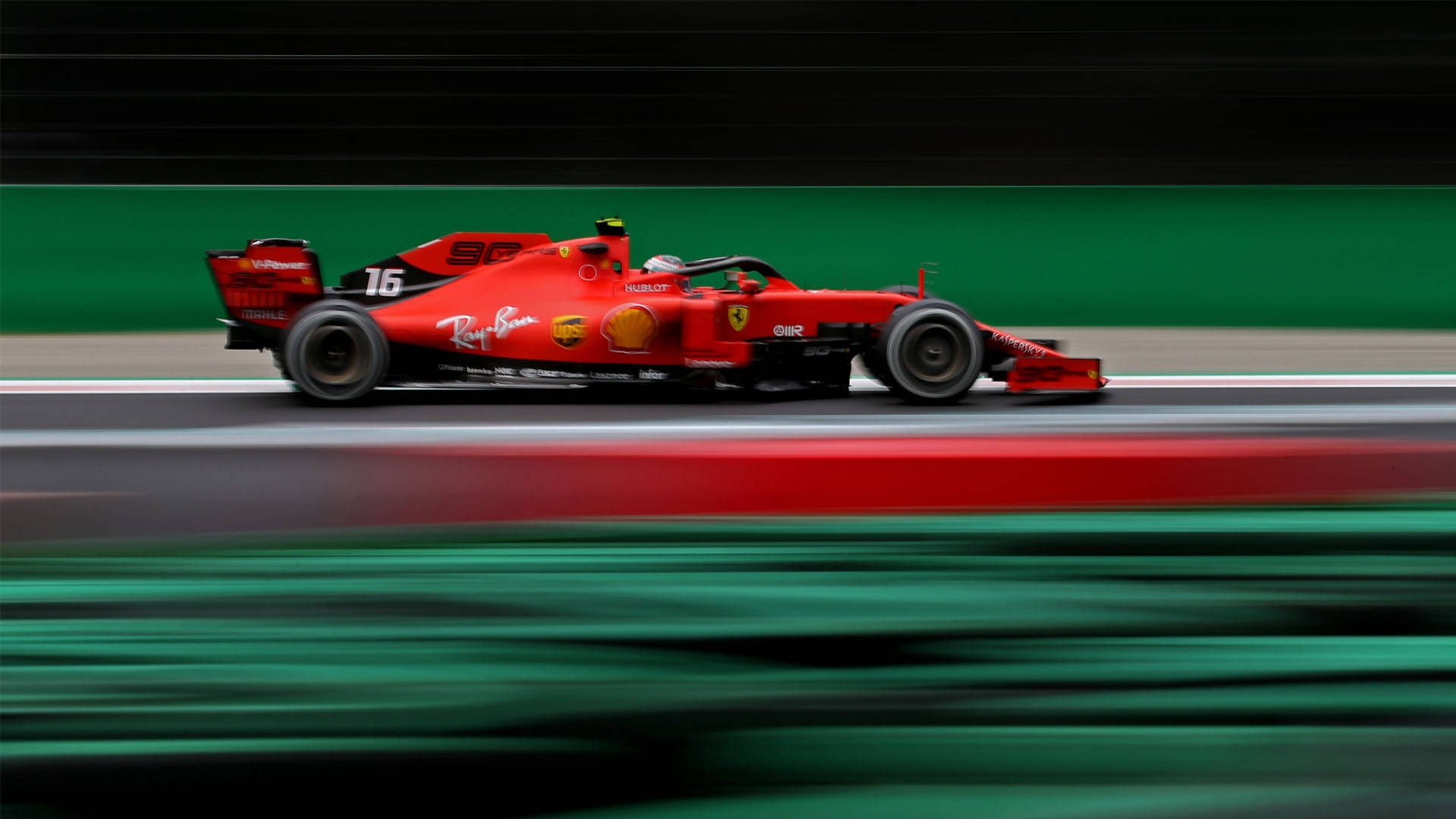 Formula One 桌布 壁紙 — 一輛紅色法拉利F1賽車在賽道上疾馳而過，帶有動態模糊效果，賽道兩旁是綠色護牆和紅色路緣石。