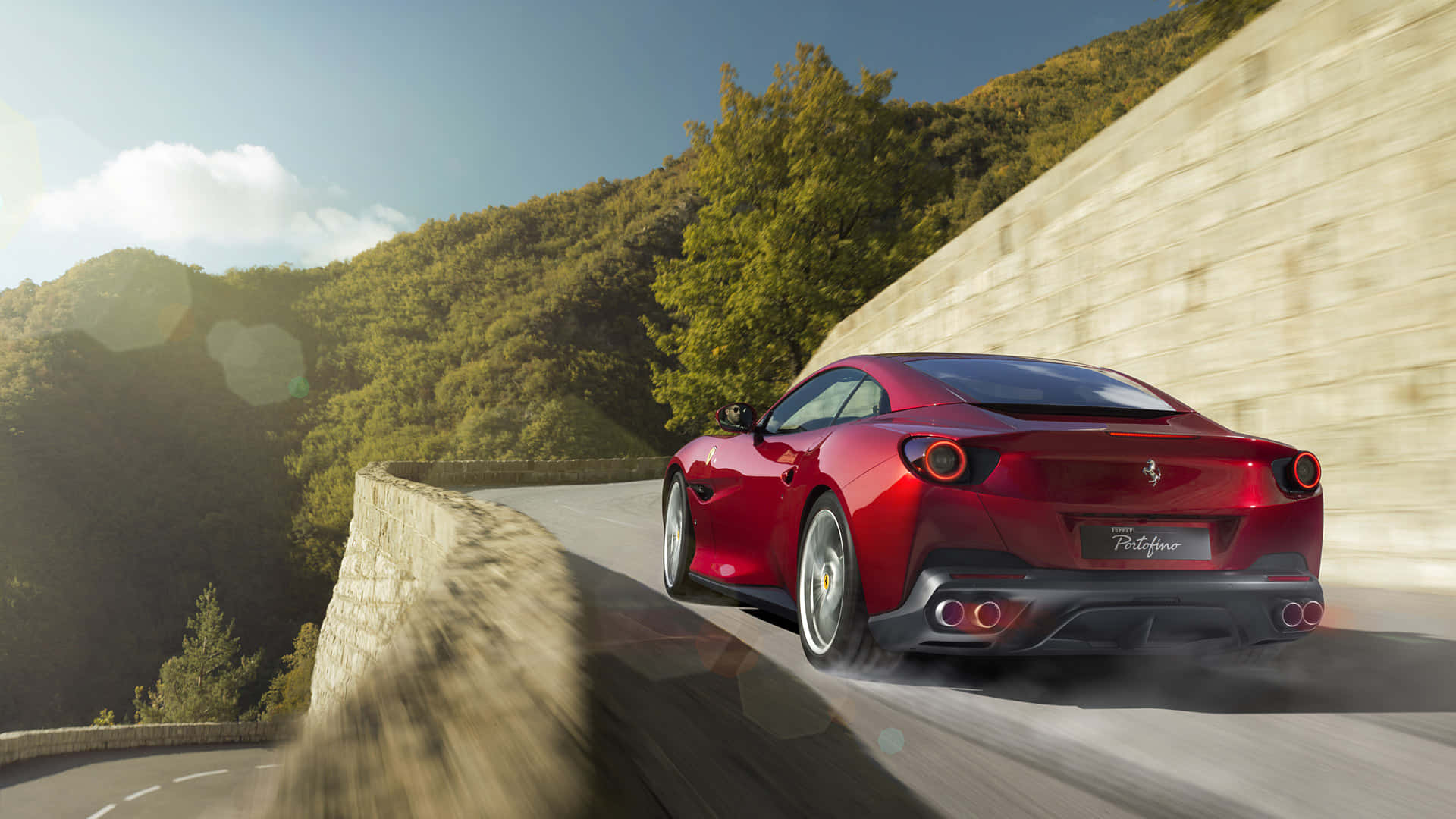 Ferrari Portofino 桌布 壁紙 — 一輛紅色法拉利Portofino在蜿蜒的山路上疾馳，兩旁是鬱鬱蔥蔥的綠色山丘和石牆，頭頂是明亮的藍天。