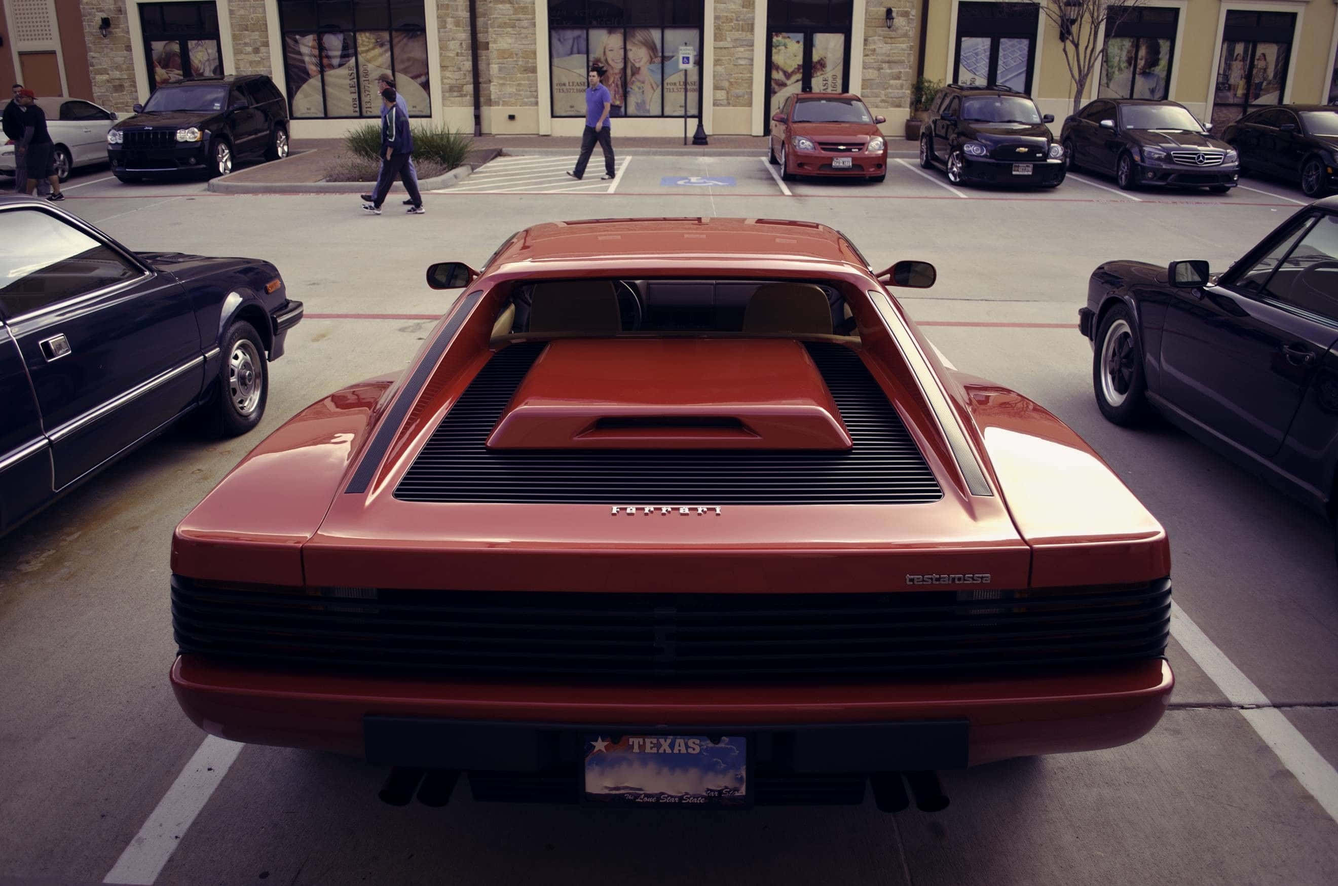 Ferrari Testarossa 桌布 壁紙 — 一輛紅色法拉利Testarossa停泊在陽光明媚的城市空地，附近有行人和車輛的後視圖。