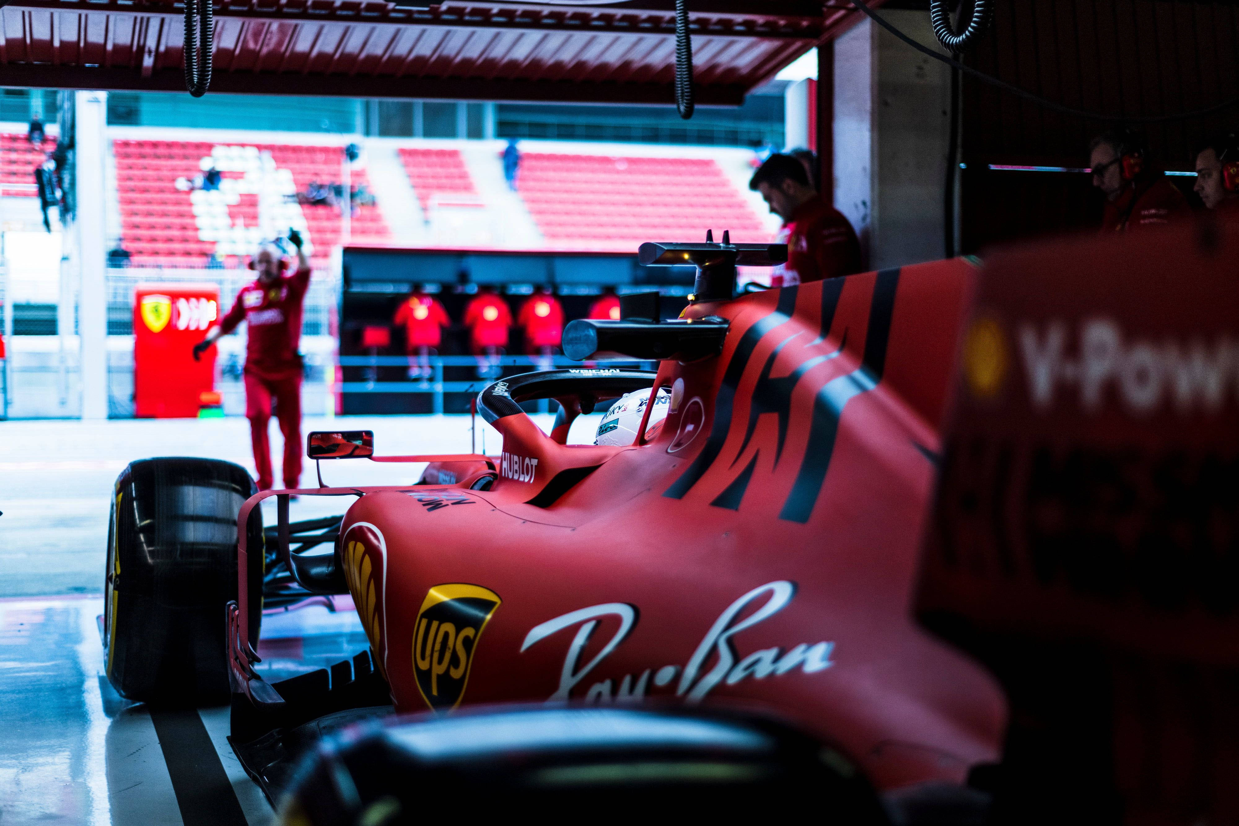 Formula One 桌布 壁紙 — 一輛紅色法拉利一級方程式賽車停在維修區車庫，身穿紅色制服的工作人員在明亮的燈光下圍繞它工作。
