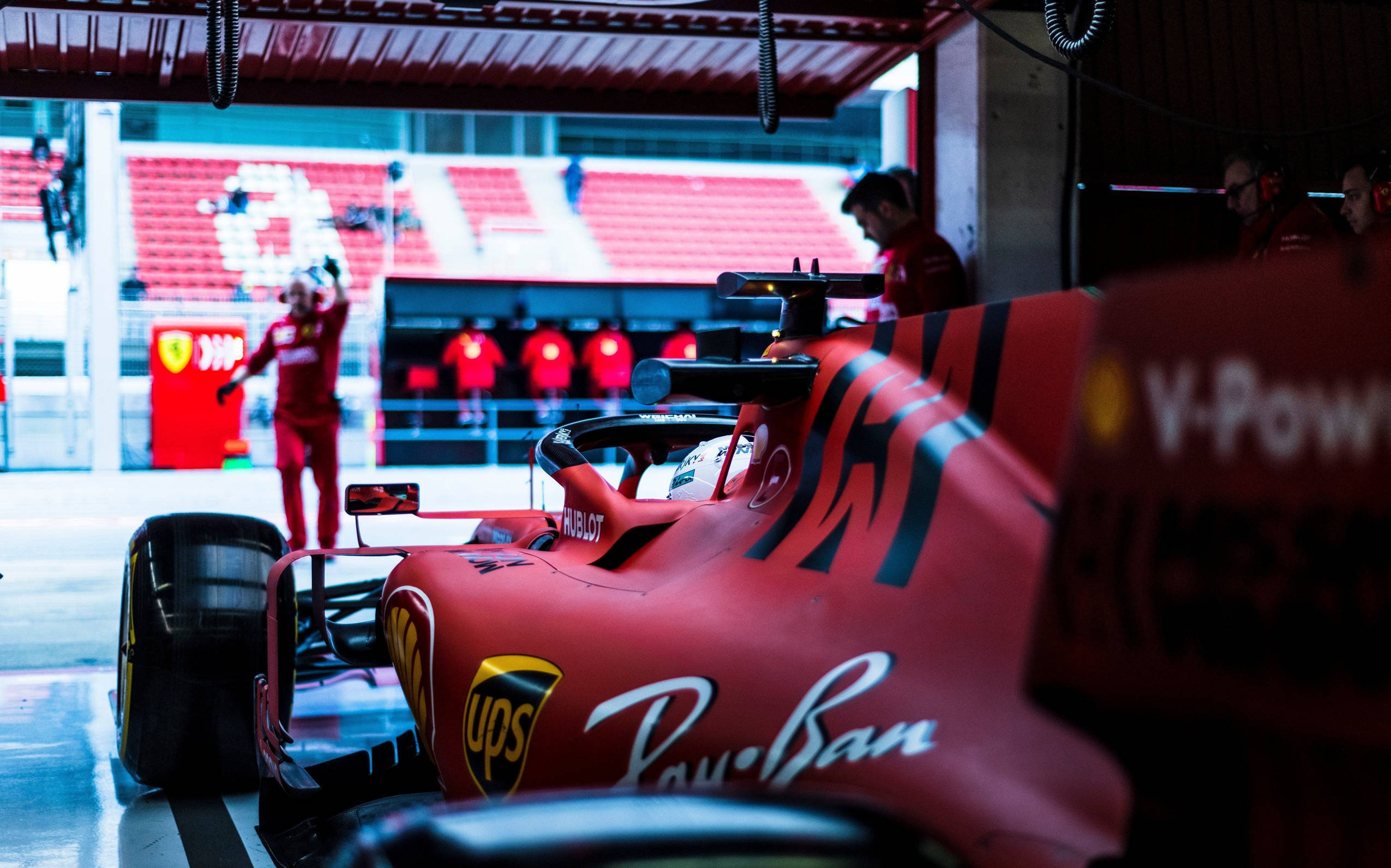 Formula 1 Desktop 桌布 壁紙 — 一輛紅色法拉利一級方程式賽車在維修站車庫內，背景可見車隊成員和鮮紅色的體育場座位。