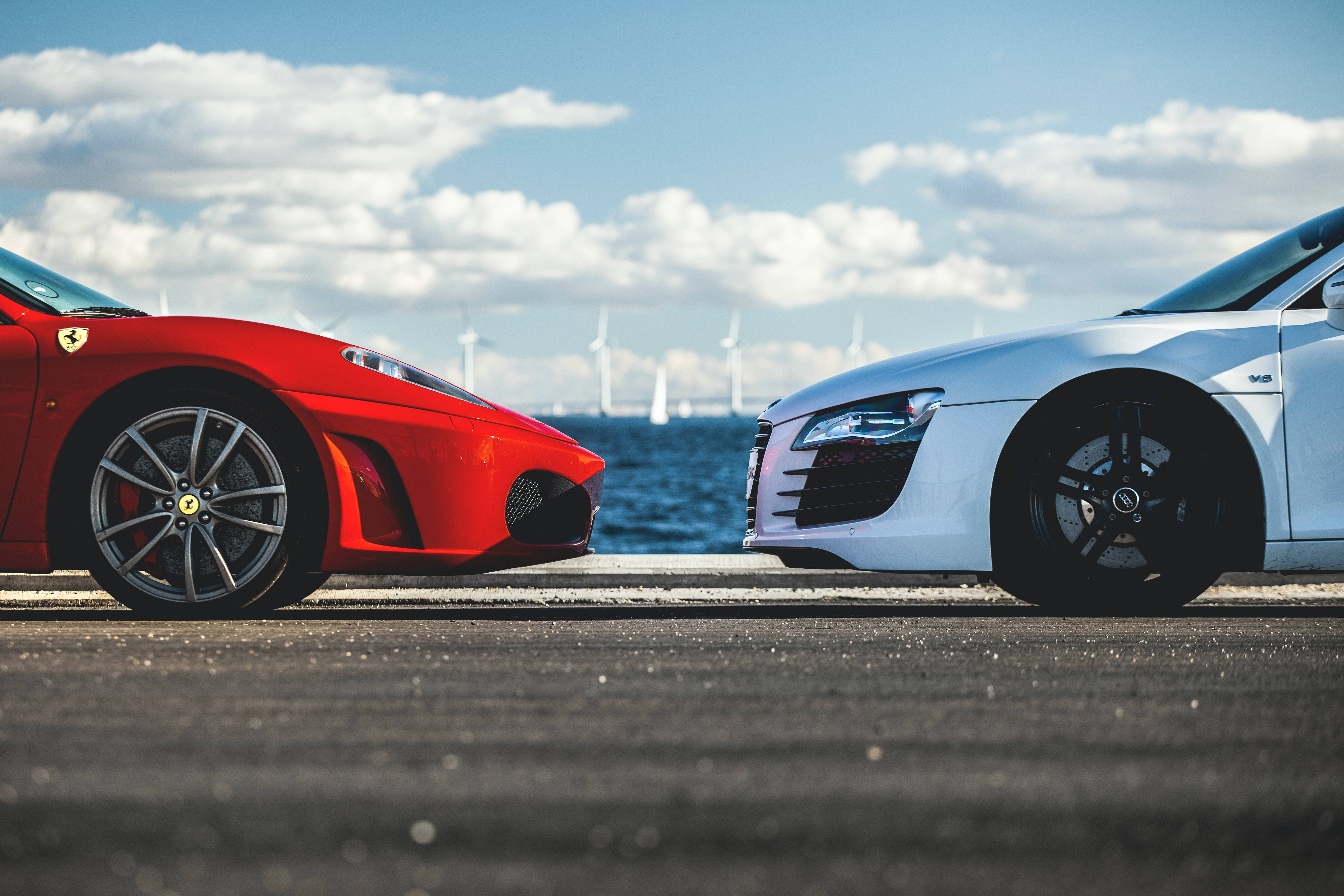 cars 桌布 壁紙 — 一輛紅色法拉利和白色奧迪R8在沿海公路上對峙，遠處有風力發電機和帆船，天空湛藍。