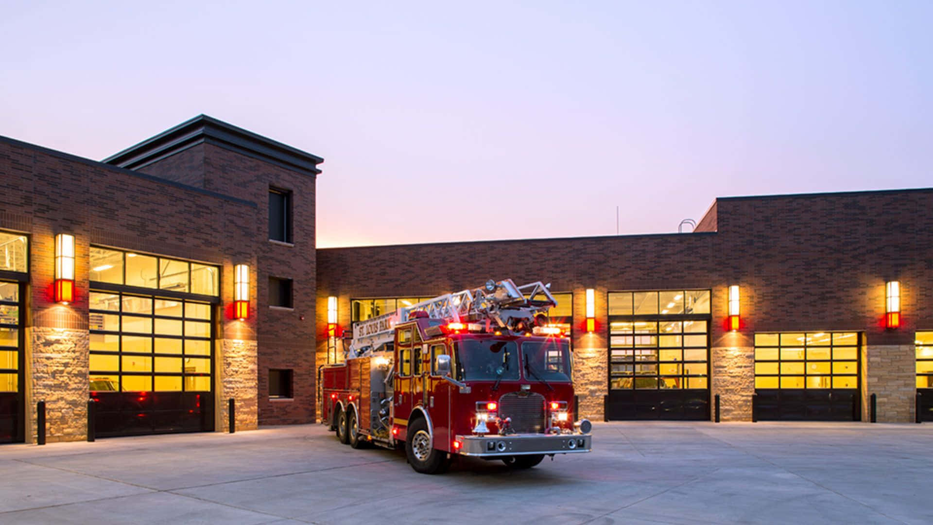 Firetruck 桌布 壁紙 — 一輛紅色消防車停放在現代磚造消防局外，黃昏時分，溫暖的室內燈光透過大窗戶照射出來。