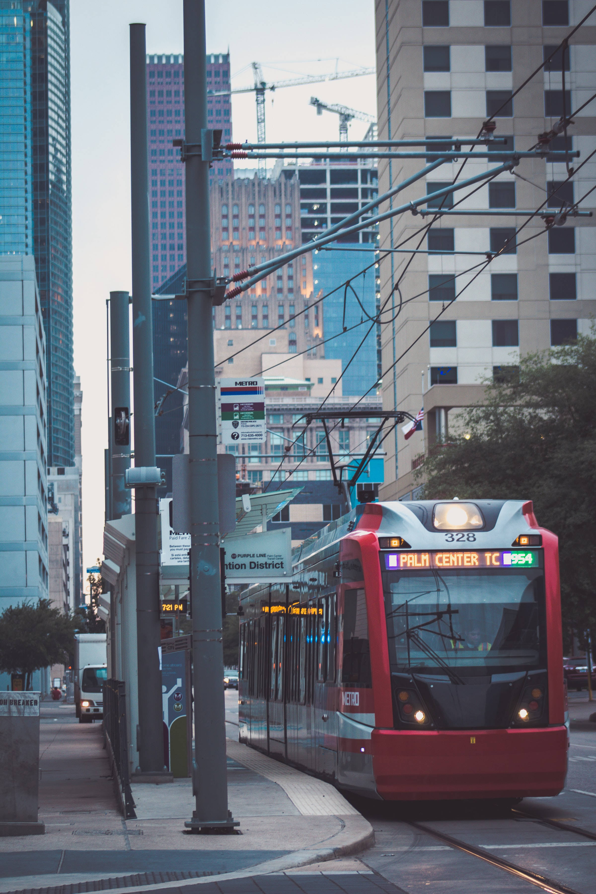 Houston 桌布 壁紙 — 一輛紅色現代電車在繁忙的城市峽谷中滑行，四周是高聳的玻璃摩天大樓和建築起重機，天空朦朧。