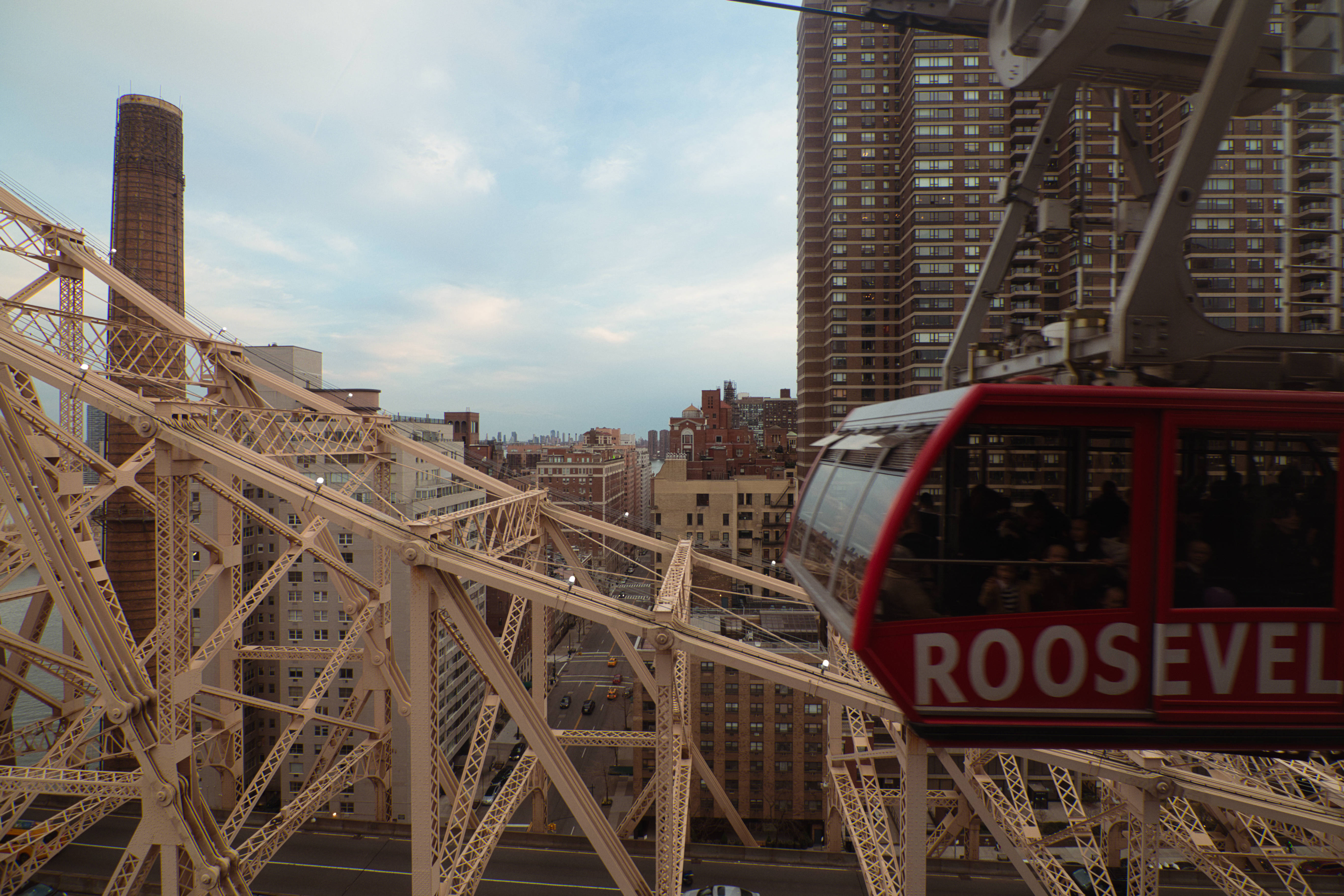 Nyc 桌布 壁紙 — 一輛紅色的羅斯福島纜車在紐約市摩天大樓的背景下，滑過一座橋樑複雜的鋼桁架，天空柔和湛藍。
