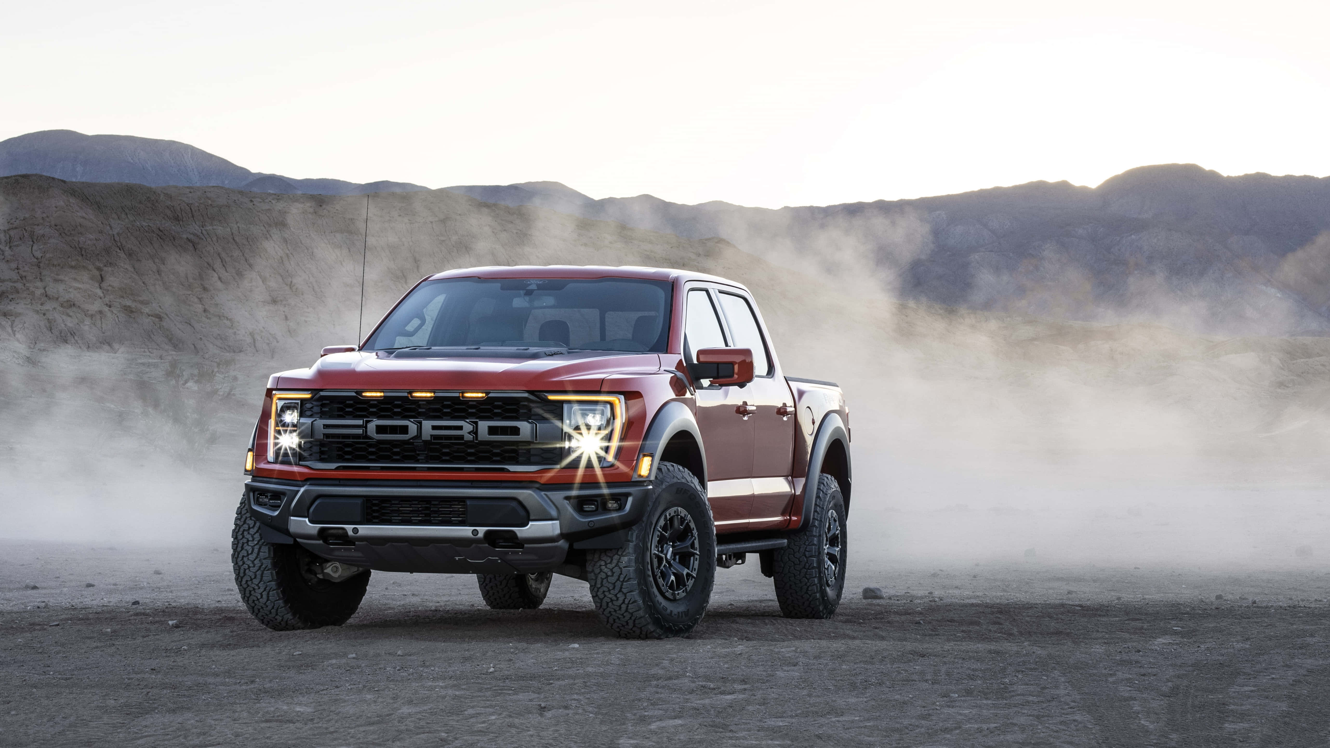 Red Car 桌布 壁紙 — 一輛紅色福特 F-150 Raptor 貨車在崎嶇的沙漠景觀中行駛，揚起塵土，正值日出時分。