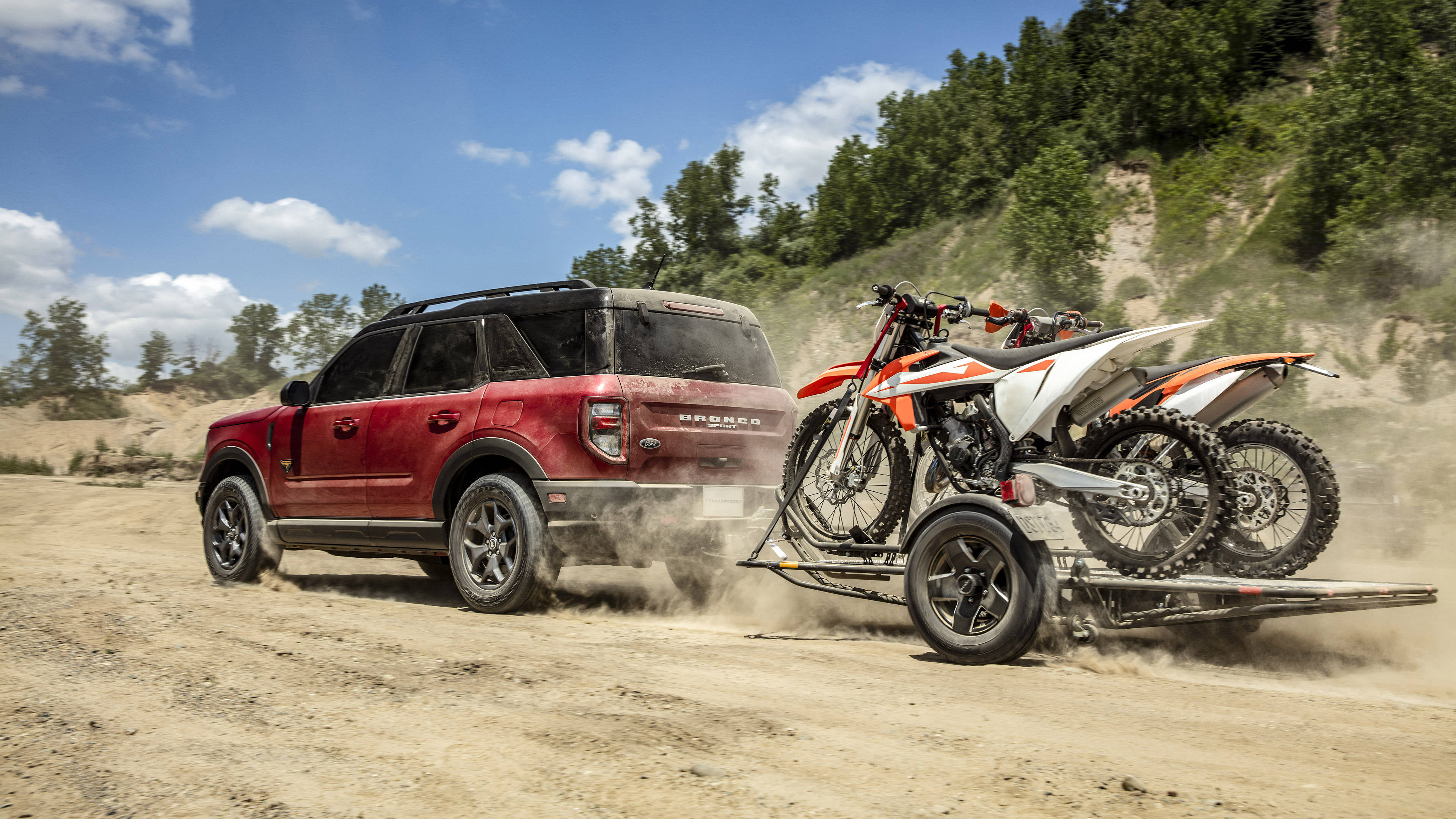 Ford Bronco 桌布 壁紙 — 一輛紅色福特Bronco SUV在陽光明媚的泥路上揚起灰塵，拖著一輛載有兩輛橙色和白色越野摩托車的拖車。