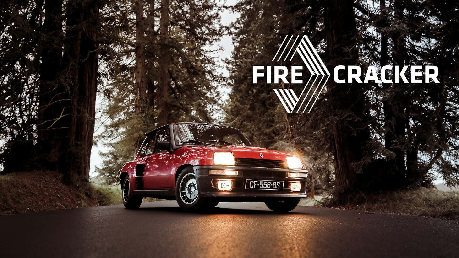 Renault 5 Turbo 桌布 壁紙 — 一輛紅色經典拉力賽車亮著車頭燈，行駛在兩旁高聳樹木的林間道路上。