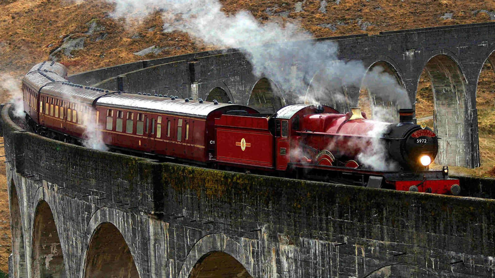 The Hogwarts Express Train 桌布 壁紙 — 一輛紅色蒸汽火車噴出煙霧，穿越崎嶇秋日山丘中的高聳石造高架橋。