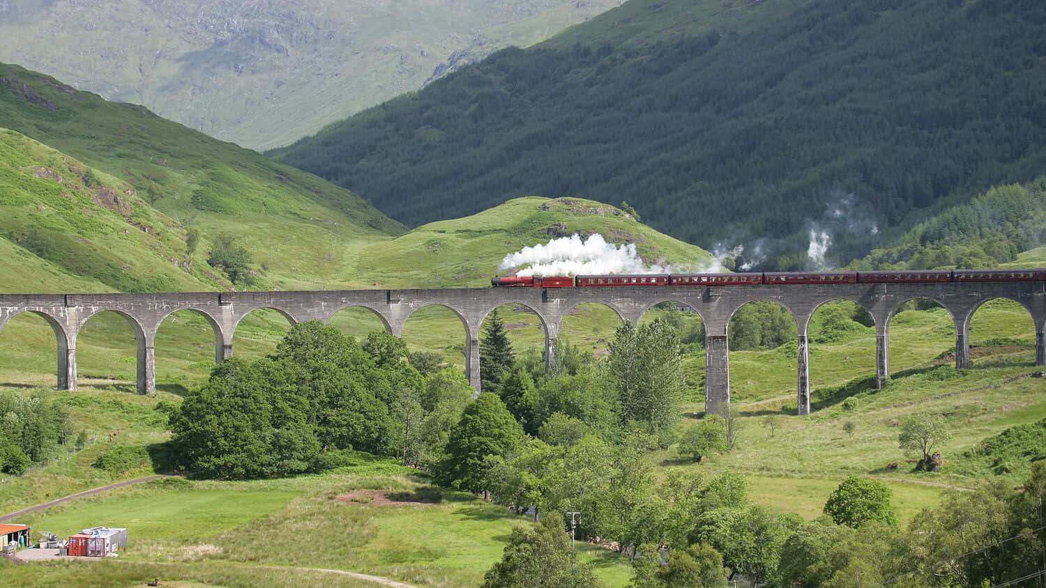 Hogwarts Express 桌布 壁紙 — 一輛紅色蒸汽火車噴出白煙，穿過石拱橋，行駛在鬱鬱蔥蔥的綠色山丘和森林之間。