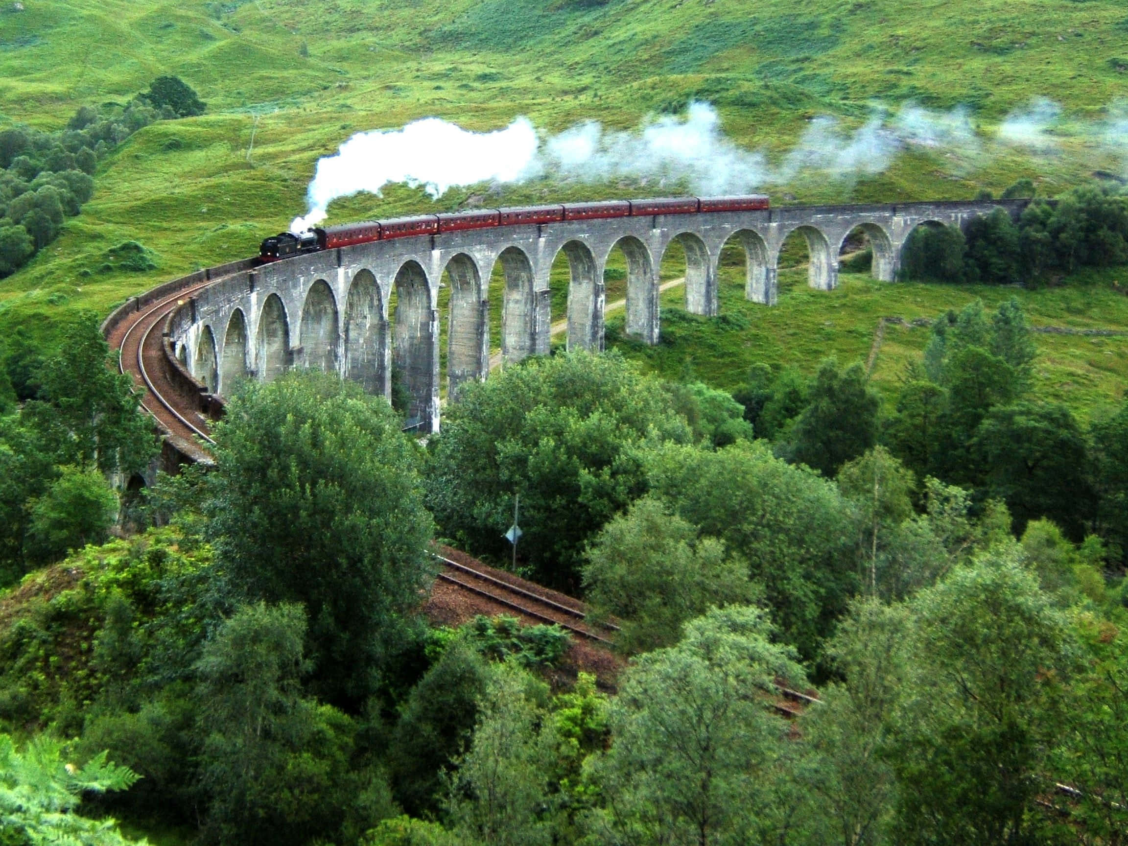 The Hogwarts Express Train 桌布 壁紙 — 一輛紅色蒸汽火車沿著彎曲的石拱橋行駛，穿過鬱鬱蔥蔥的綠色山丘和茂密的森林，噴出白色煙霧。