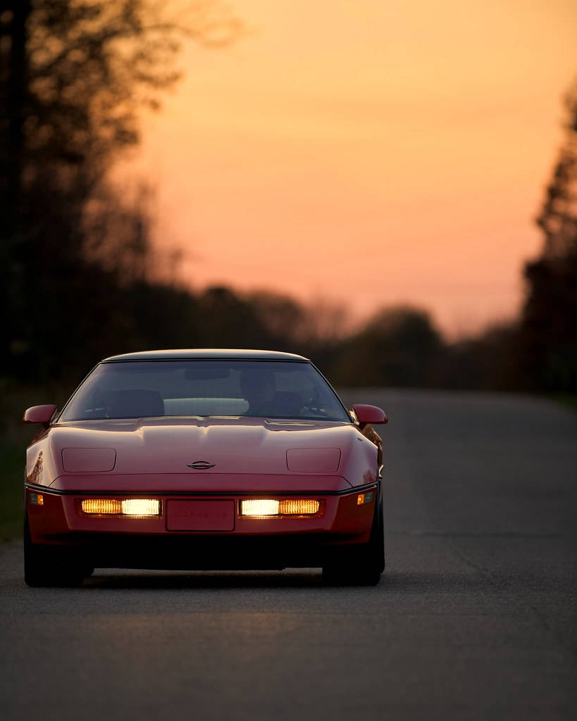 corvette 桌布 壁紙 — 一輛紅色跑車在日落時分，溫暖橙色天空下，在寧靜的道路上亮著車頭燈向前行駛。
