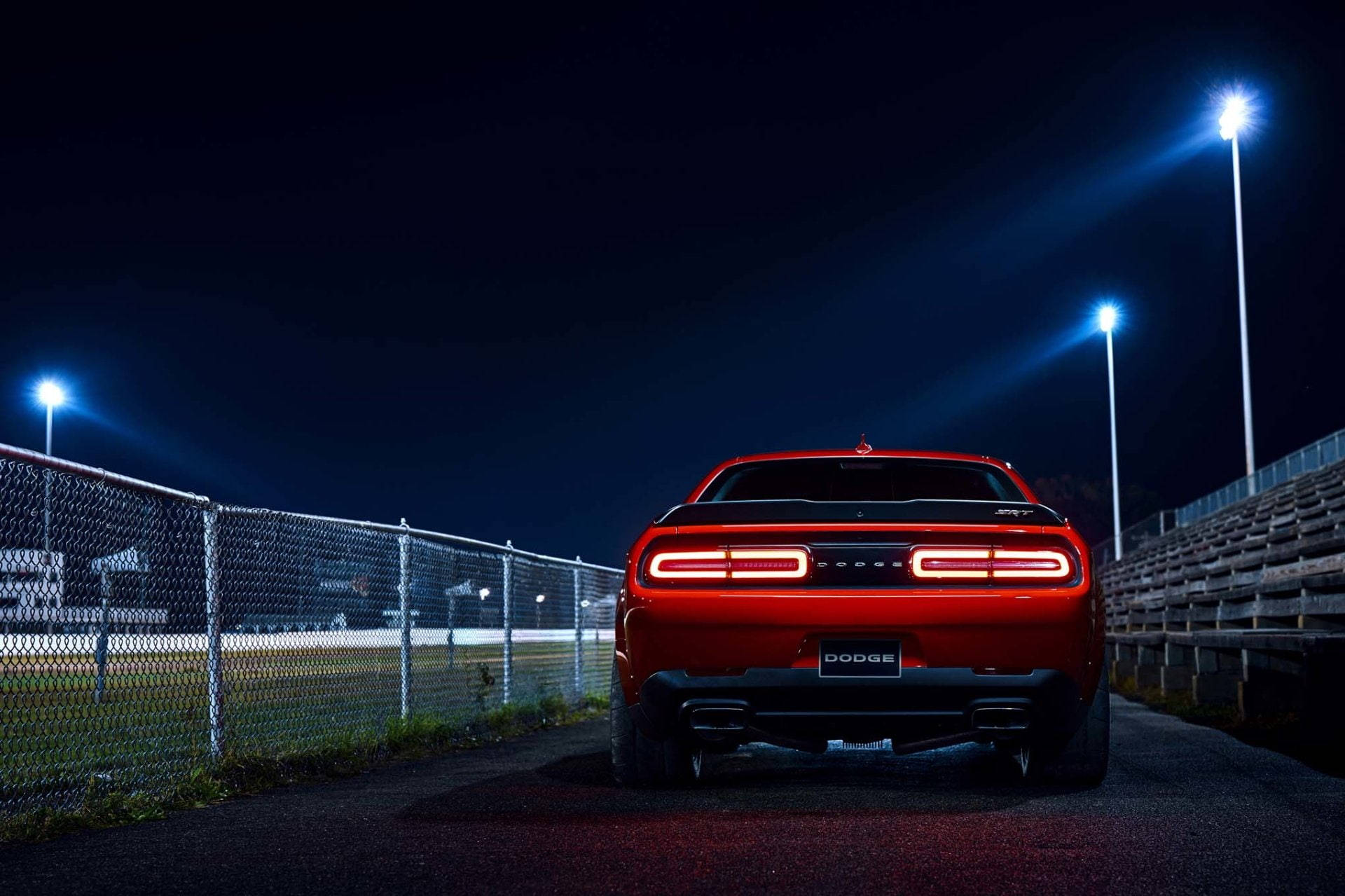 Mopar 桌布 壁紙 — 一輛紅色道奇挑戰者在夜間停放在明亮的體育場燈光下，背景有鐵絲網圍欄和看台。