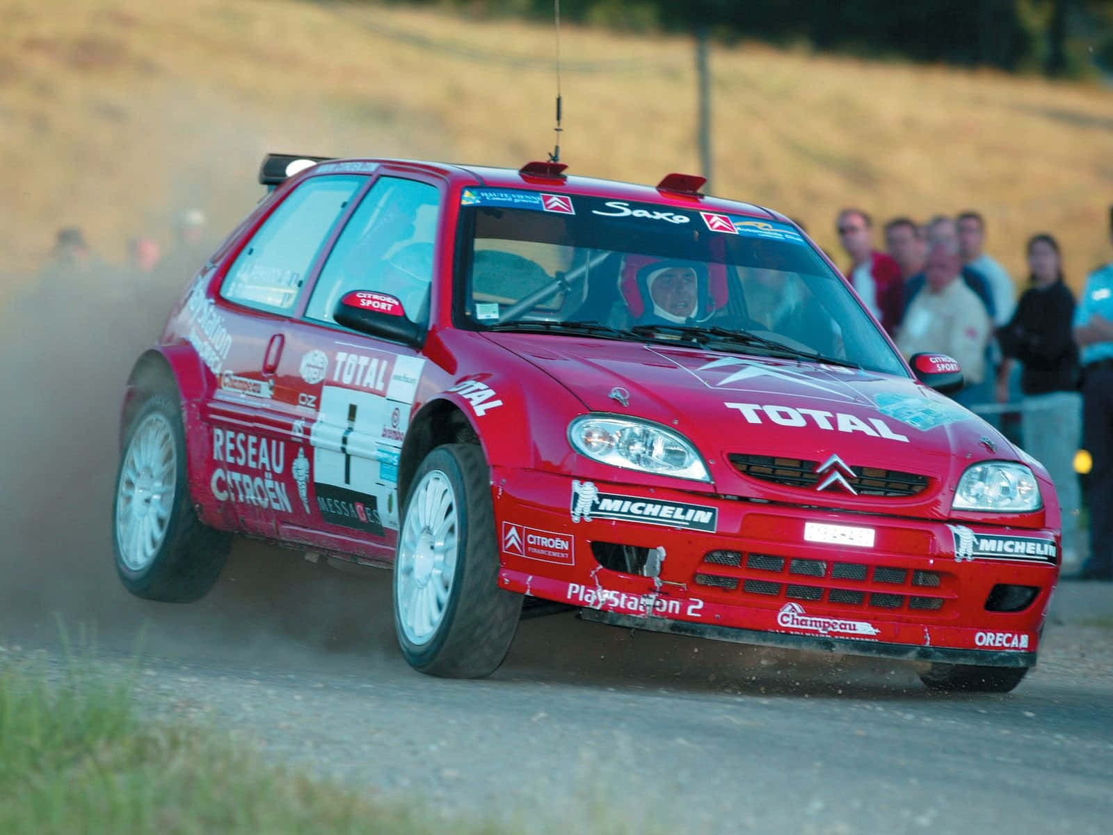 Citroen C2 Super 1600 桌布 壁紙 — 一輛紅色雪鐵龍拉力賽車在土路上疾馳，揚起灰塵，觀眾在旁觀看。
