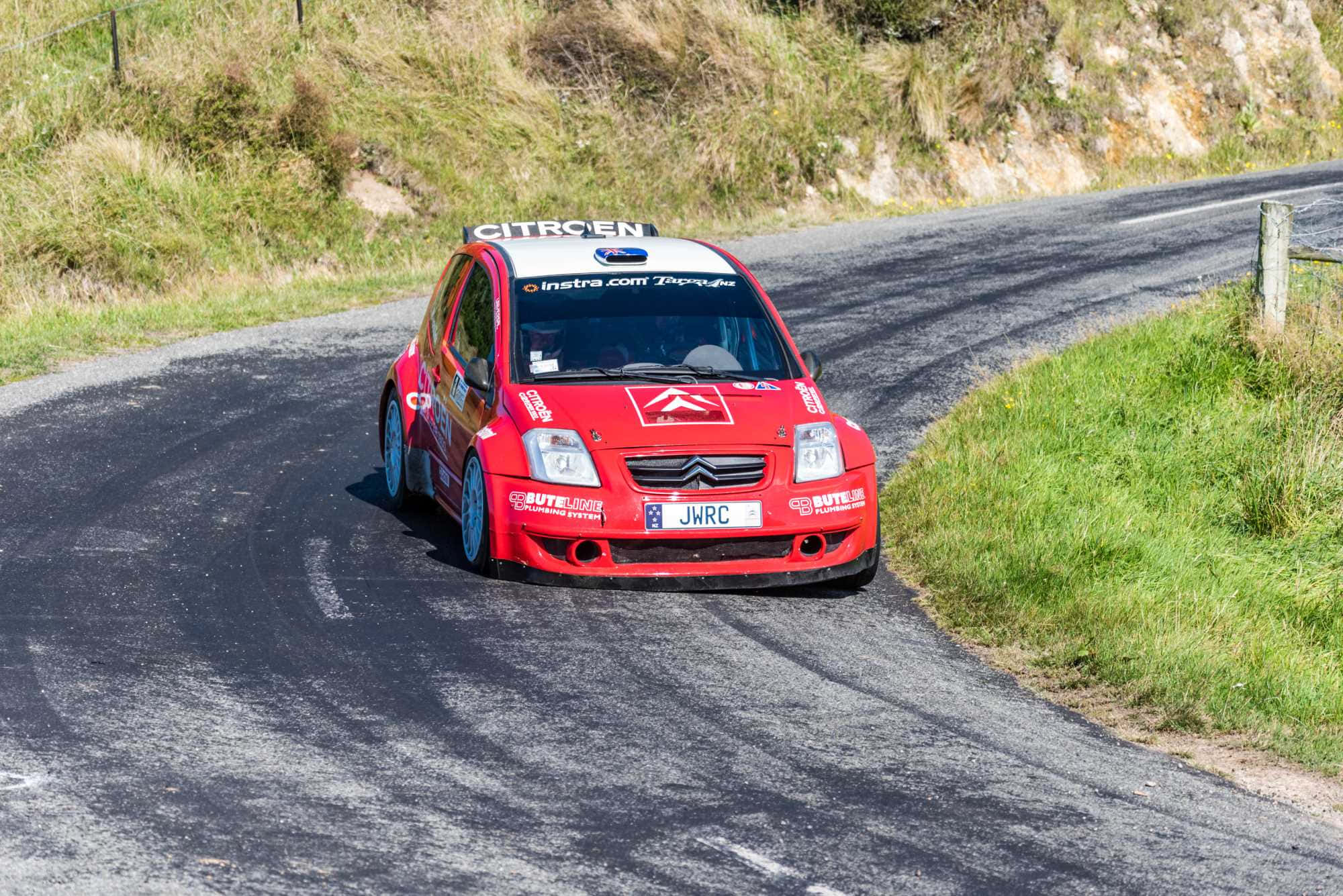 Citroen C2 Super 1600 桌布 壁紙 — 一輛紅色雪鐵龍拉力賽車在蜿蜒的鄉村瀝青路上高速駛過一個急彎，路邊是長滿青草的山丘。
