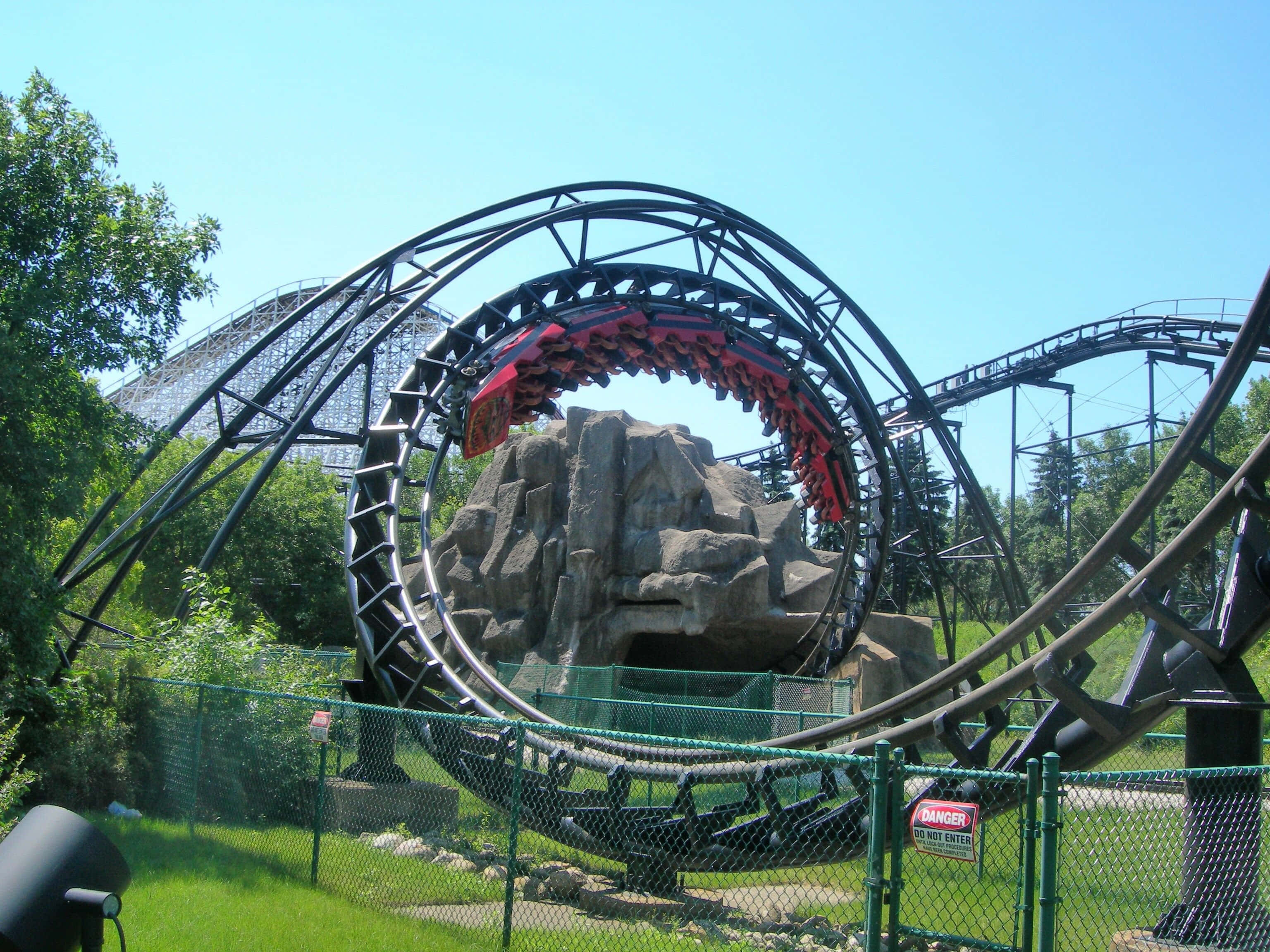 Roller Coaster 桌布 壁紙 — 一輛紅色雲霄飛車在晴朗的藍天下，穿過高聳的黑色環圈，周圍環繞著綠樹和岩石地形。