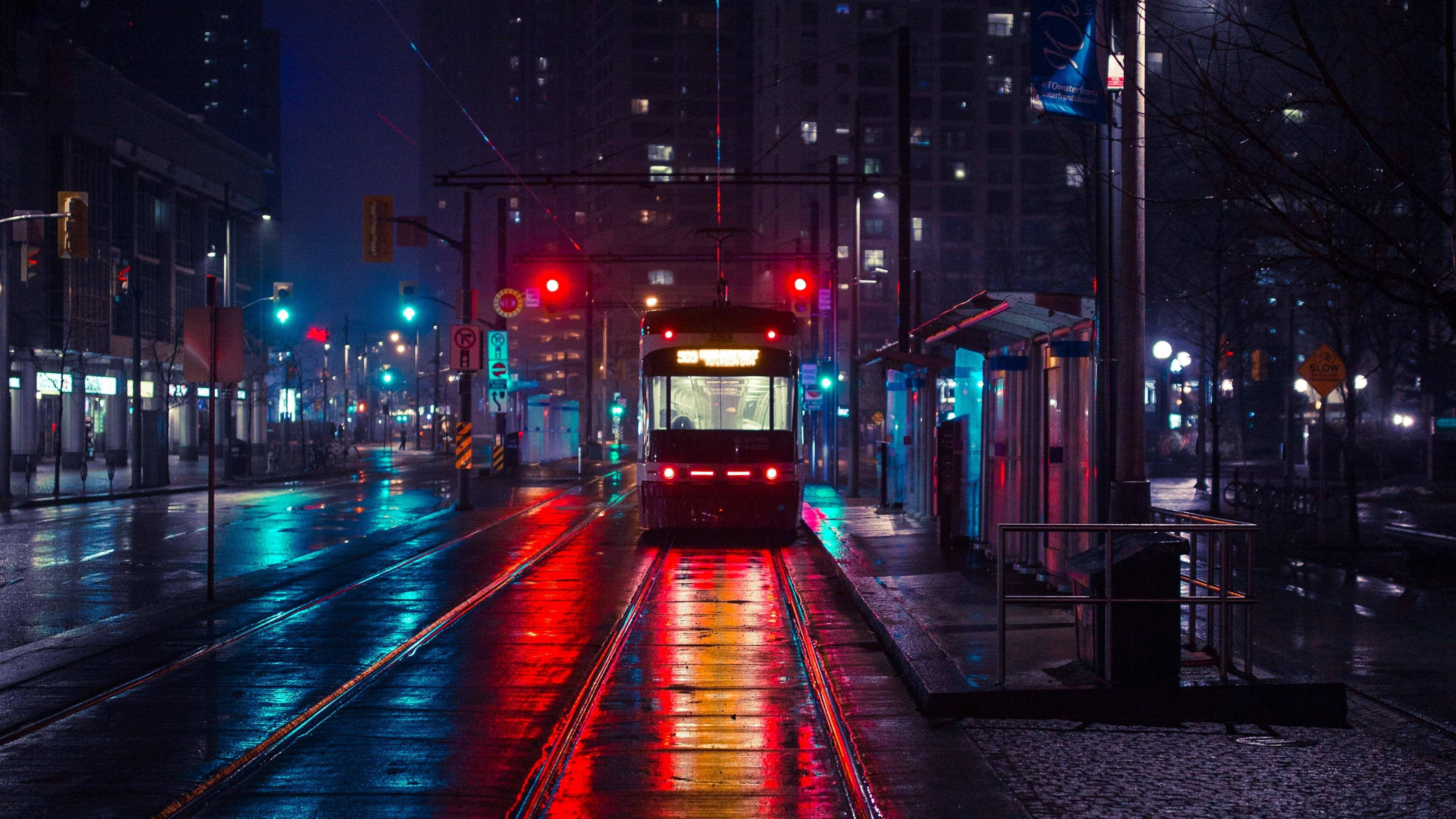 電力 桌布 壁紙 — 一輛紅色電車在雨濕的城市夜晚中滑行，其燈光在高聳建築物下的濕滑路軌和路面上生動地反射。