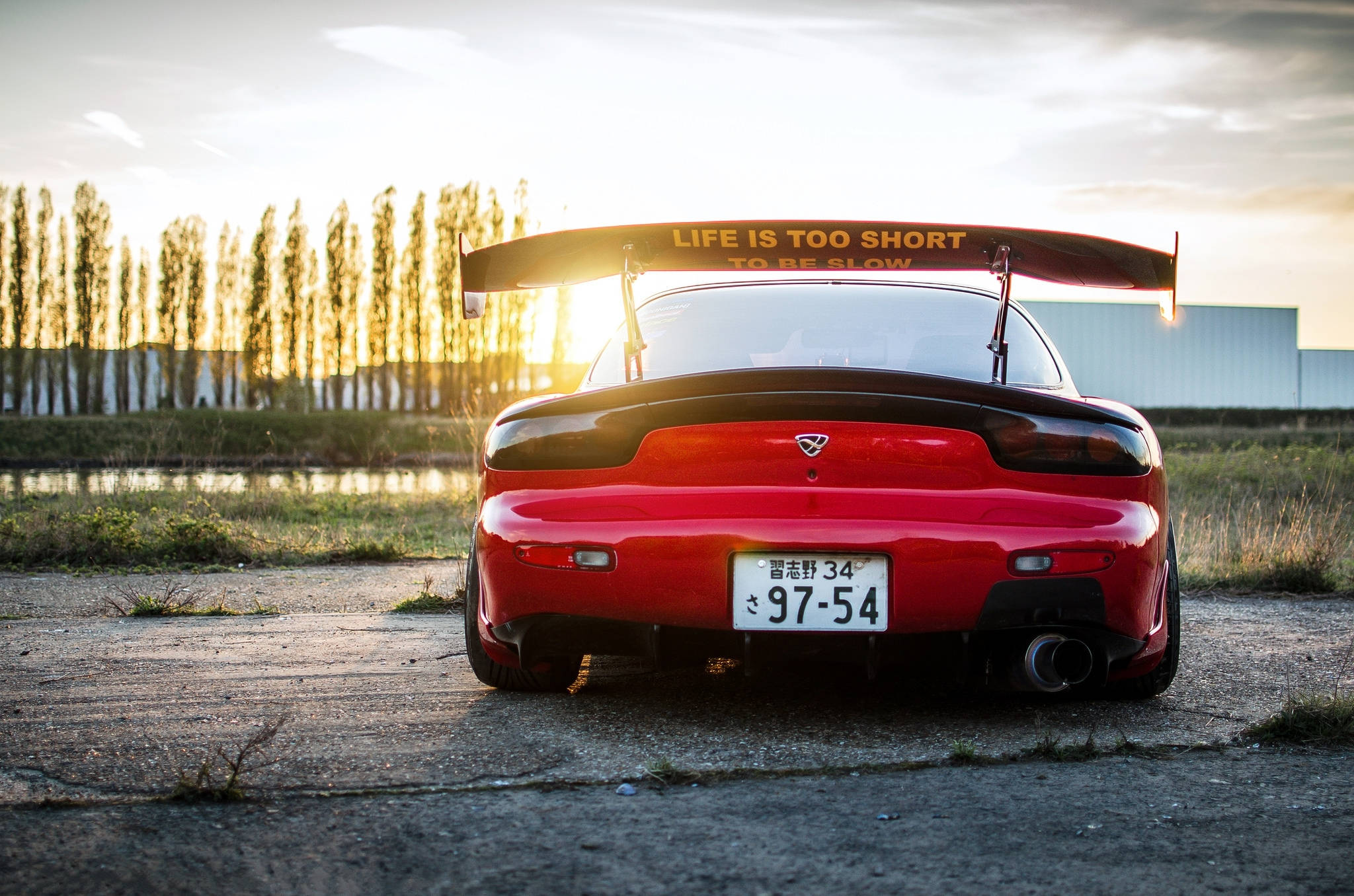 Mazda 桌布 壁紙 — 一輛紅色馬自達RX-7，帶有大型尾翼和標語「LIFE IS TOO SHORT TO BE SLOW」，停泊在日落時分龜裂的柏油路上。