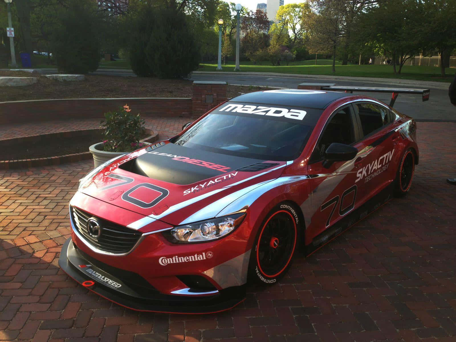 Mazda 6 桌布 壁紙 — 一輛紅色馬自達賽車，號碼70，停在磚砌廣場上，周圍有樹木和城市建築。