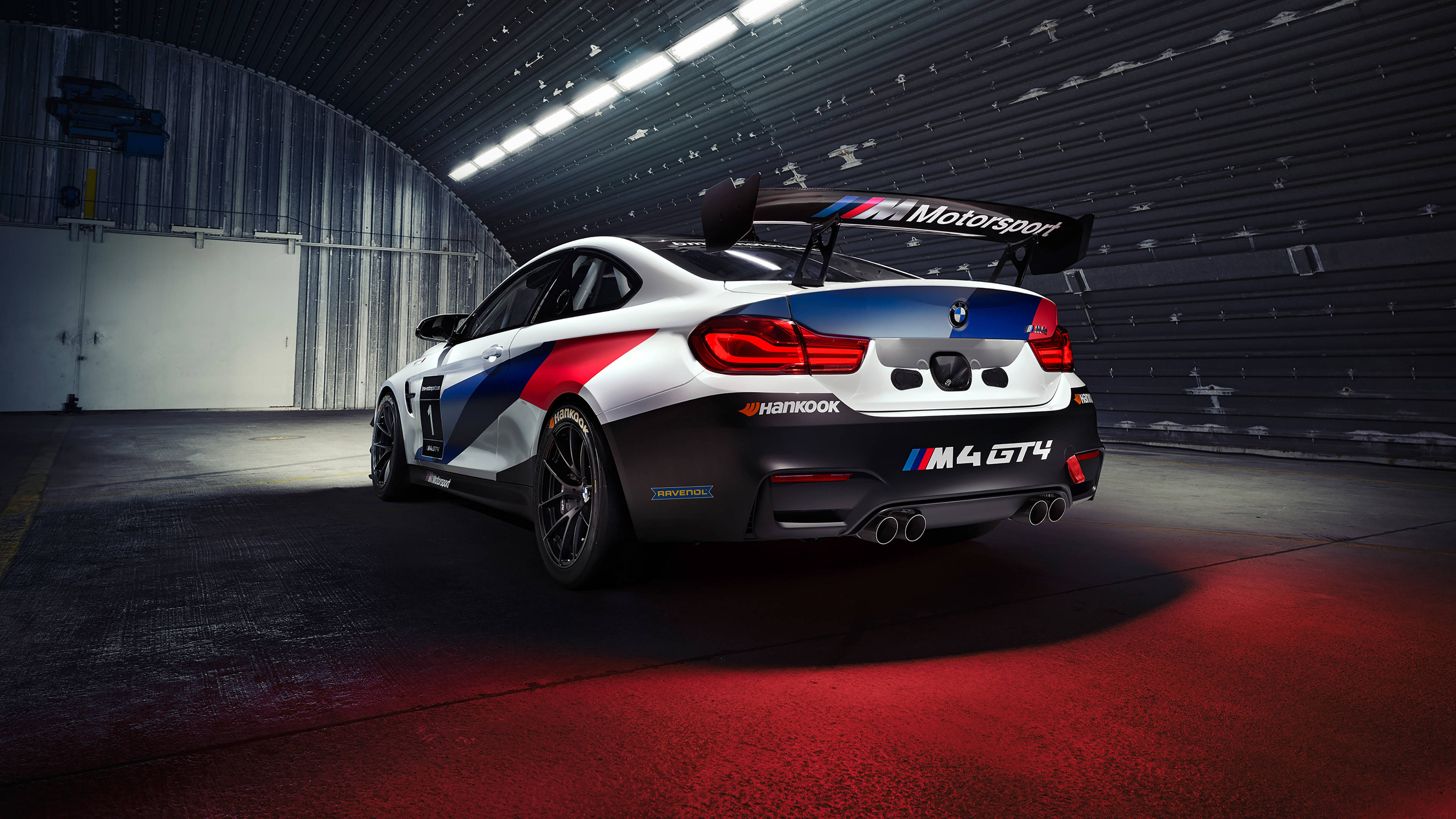 BMW 動圖 桌布 壁紙 — 一輛紅藍白塗裝的BMW M4 GT4賽車停放在光線昏暗、帶有紅色地板照明的車庫中。